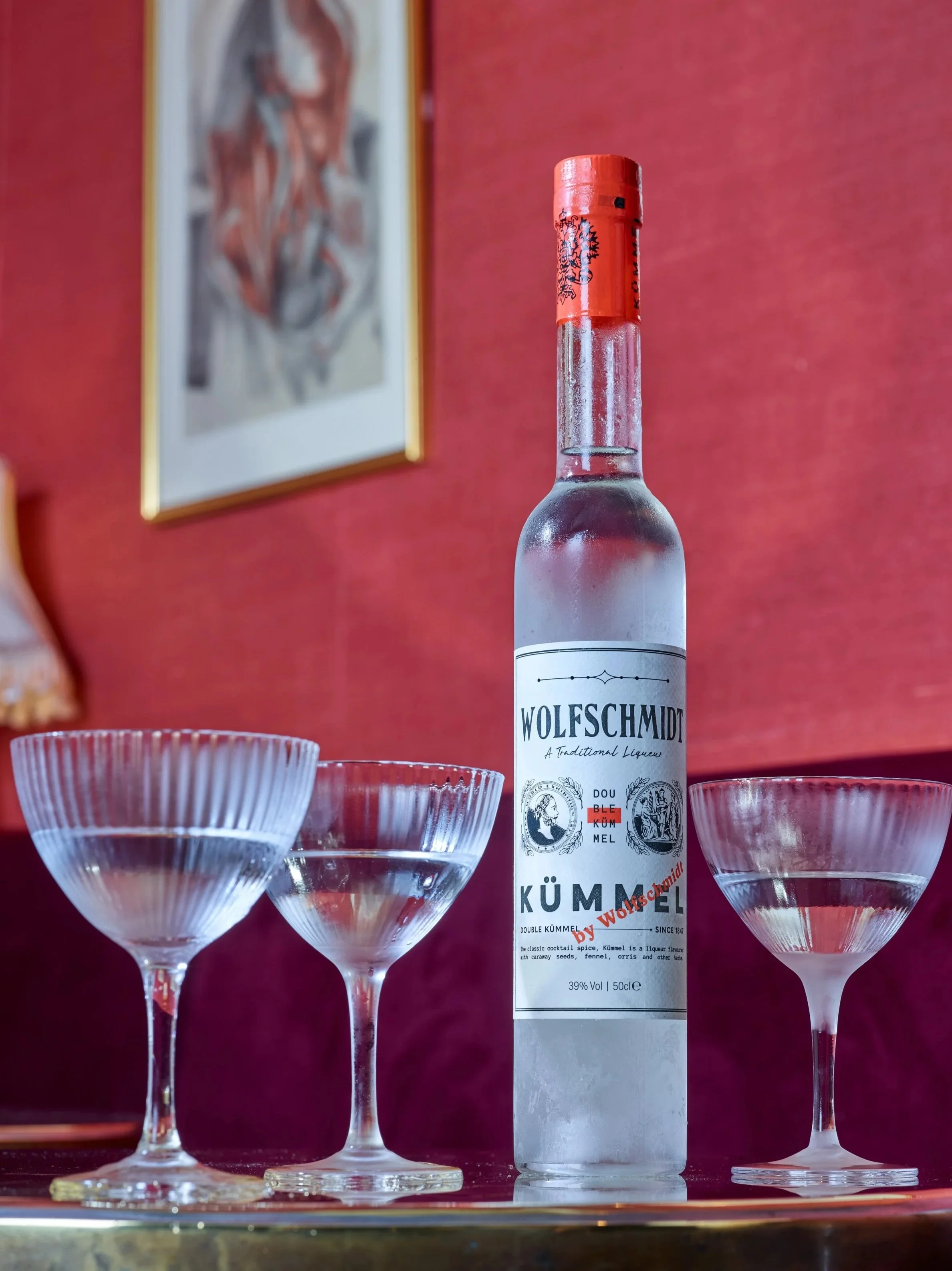 A bottle of Wolfschmidt Kumme with a red cap, surrounded by four empty, stemmed cocktail glasses, on a reflective surface with a red background and a blurry framed picture on the wall.
