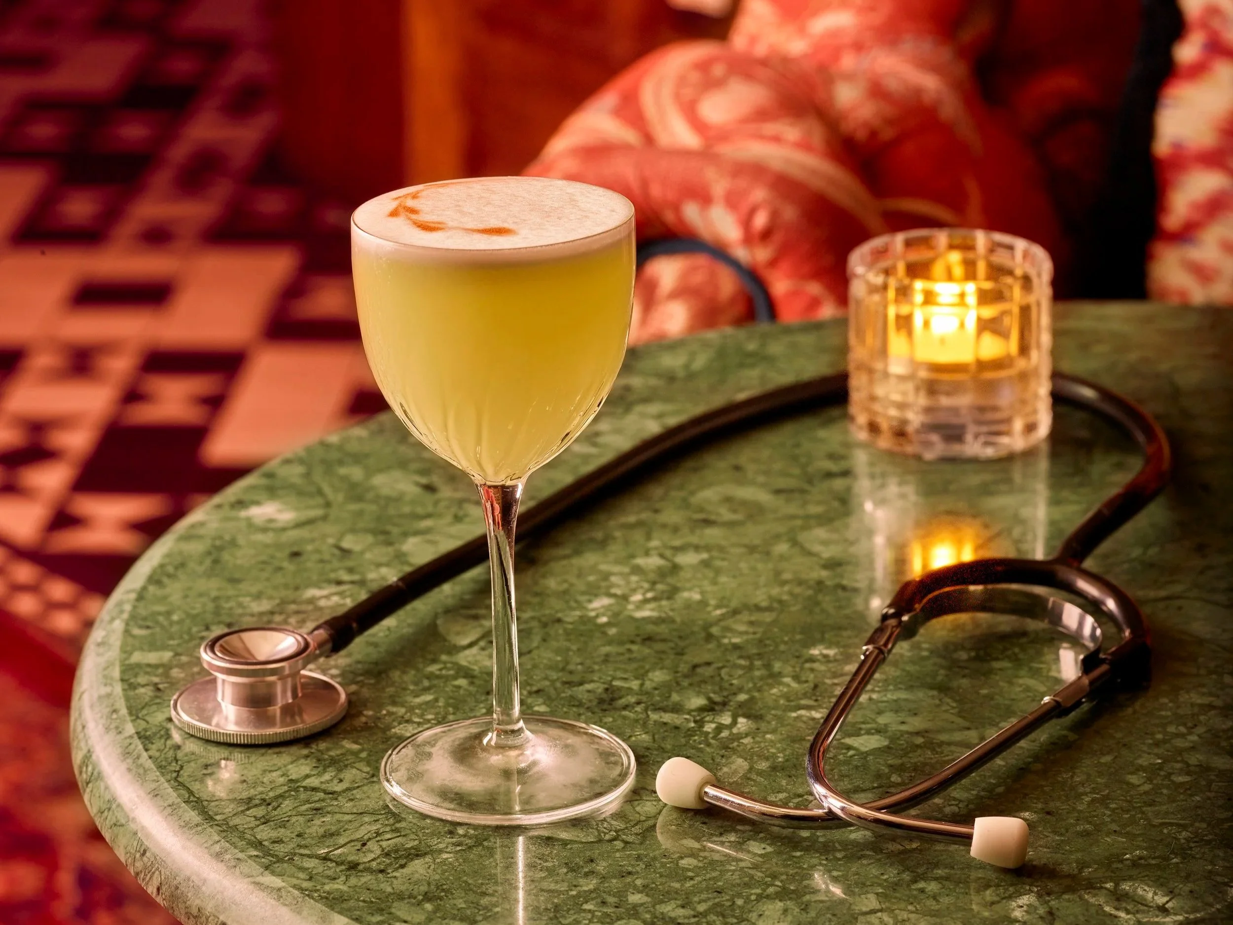 A green marble table with a glass of Wolfschmidt Kümmels recommended cocktail, a yellow-green drink with foam on top, a stethoscope, a stethoscope earpiece, and a small candle holder with a lit candle.