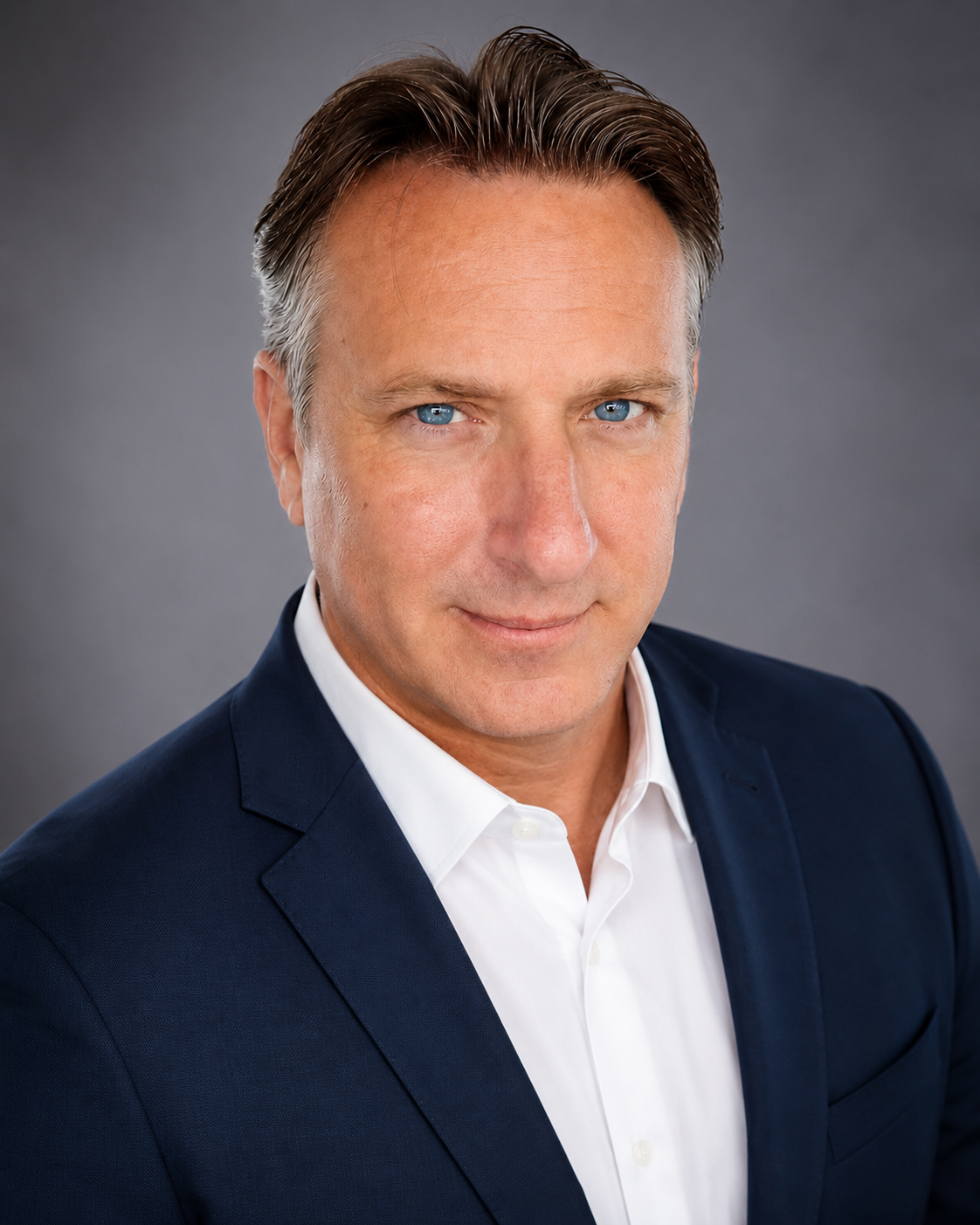 Close-up portrait of a middle-aged man with short brown hair and blue eyes, dressed in a dark blue suit and white shirt, smiling slightly against a grey background.