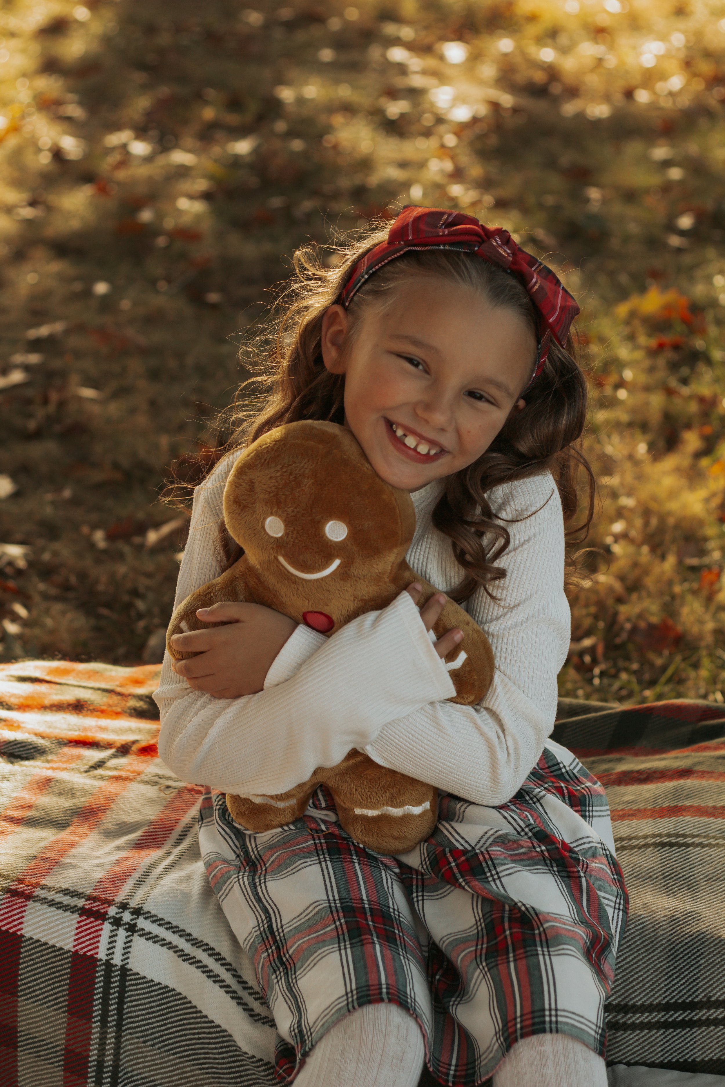 A young girl with curly hair, wearing a red plaid headband and a white sweater, sitting on a blanket outdoors during autumn. She is smiling and hugging a gingerbread man plush toy.