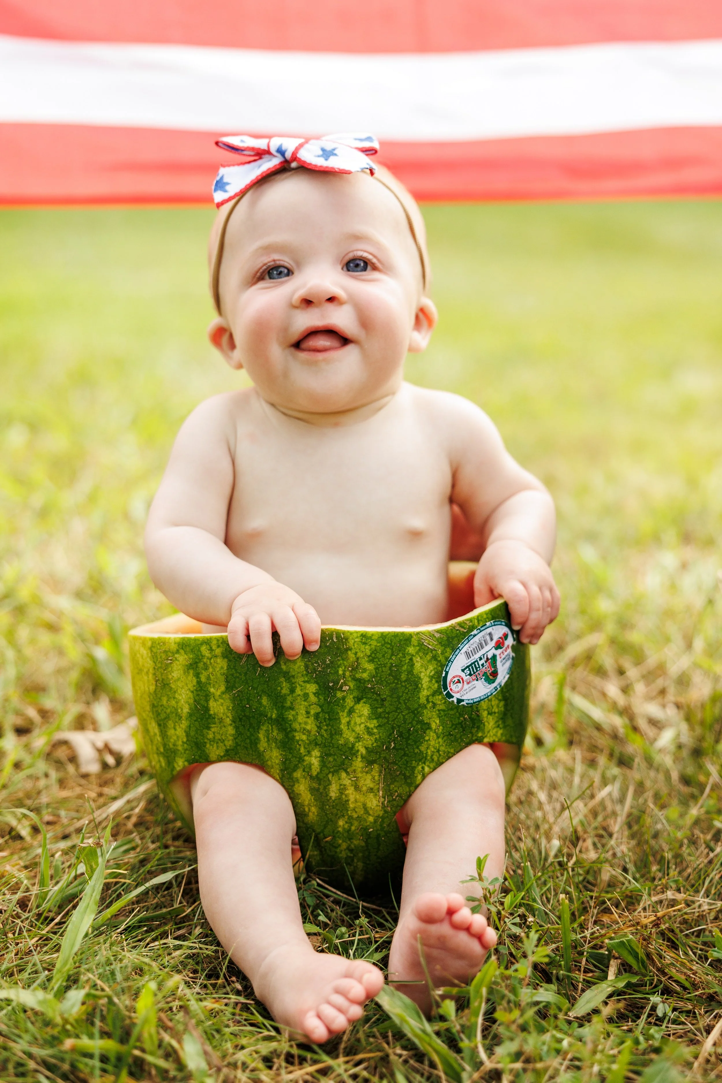 A baby sitting in a watermelon half on a grassy outdoor area, wearing a patriotic headband with stars and stripes.