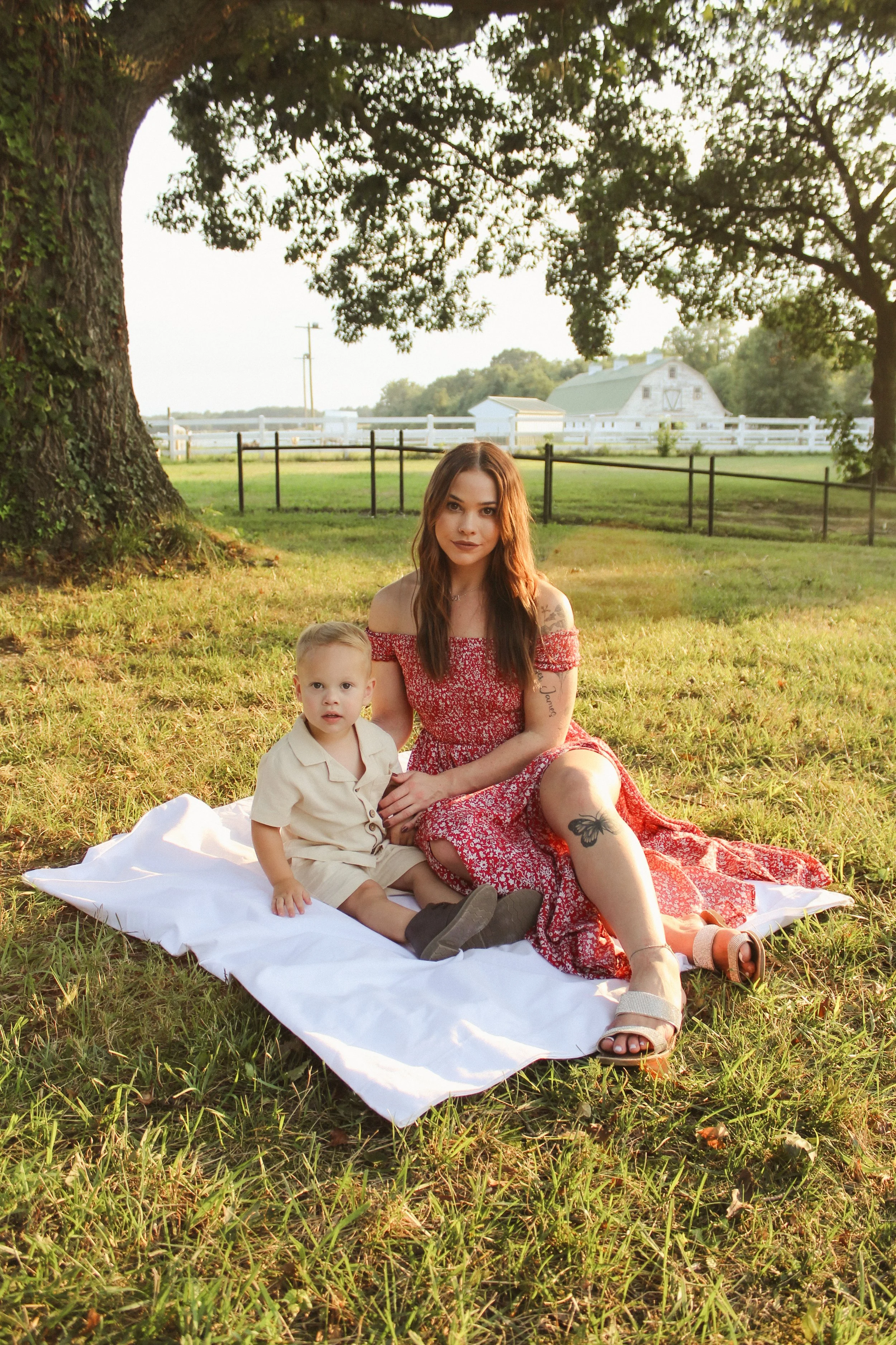 A woman and a young boy sitting on a white blanket under a large tree in a grassy field. The woman is wearing a red patterned dress, and the boy is in a beige outfit. In the background, there is a white fence, a barn, and an open farm landscape.