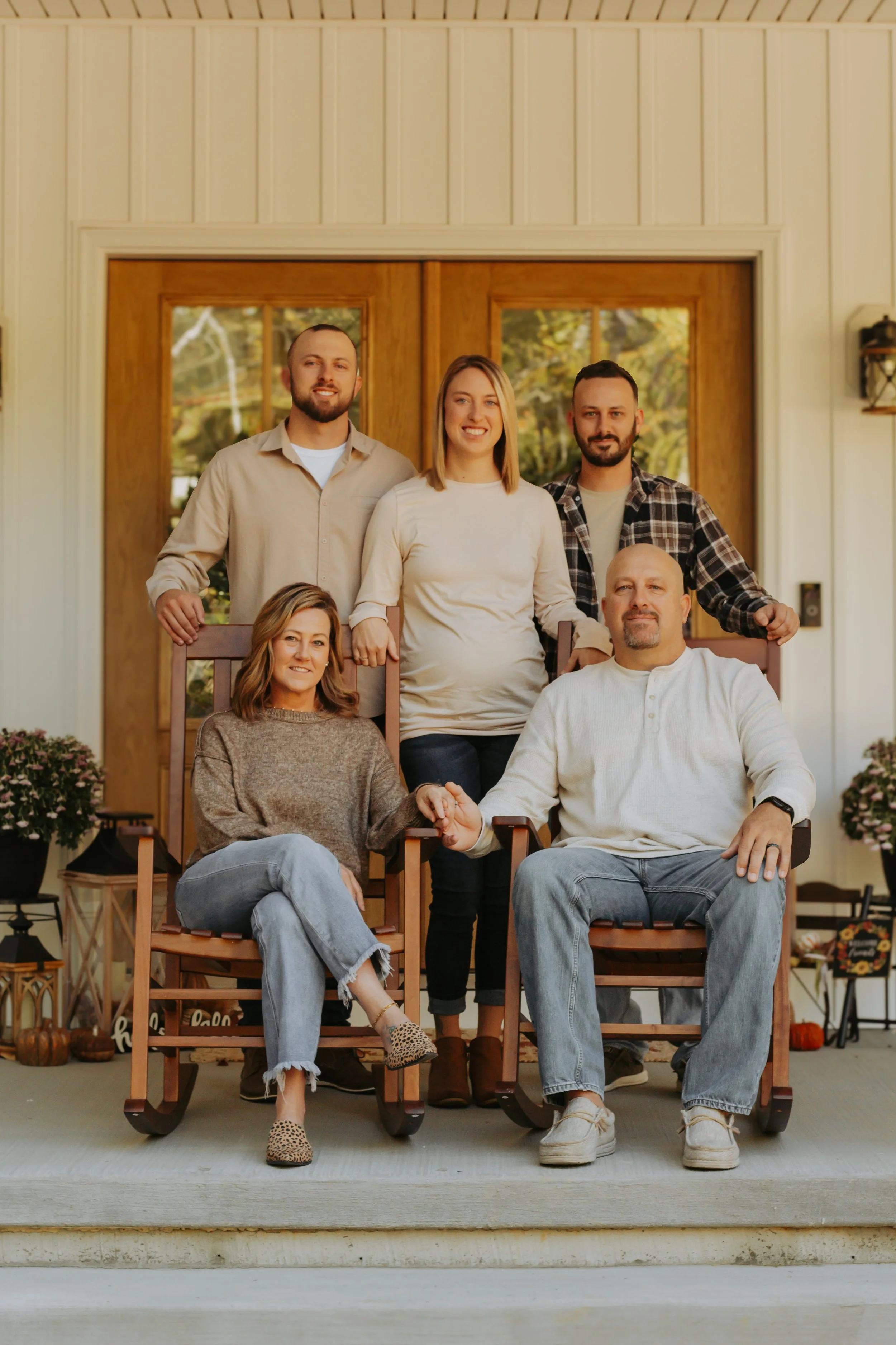 Six people, three men and three women, posing on a porch with wooden and glass double doors behind them. Two women are seated in wooden rocking chairs, holding hands, while the three men and one woman stand behind them. The scene is warmly lit with autumn decorations on the porch.
