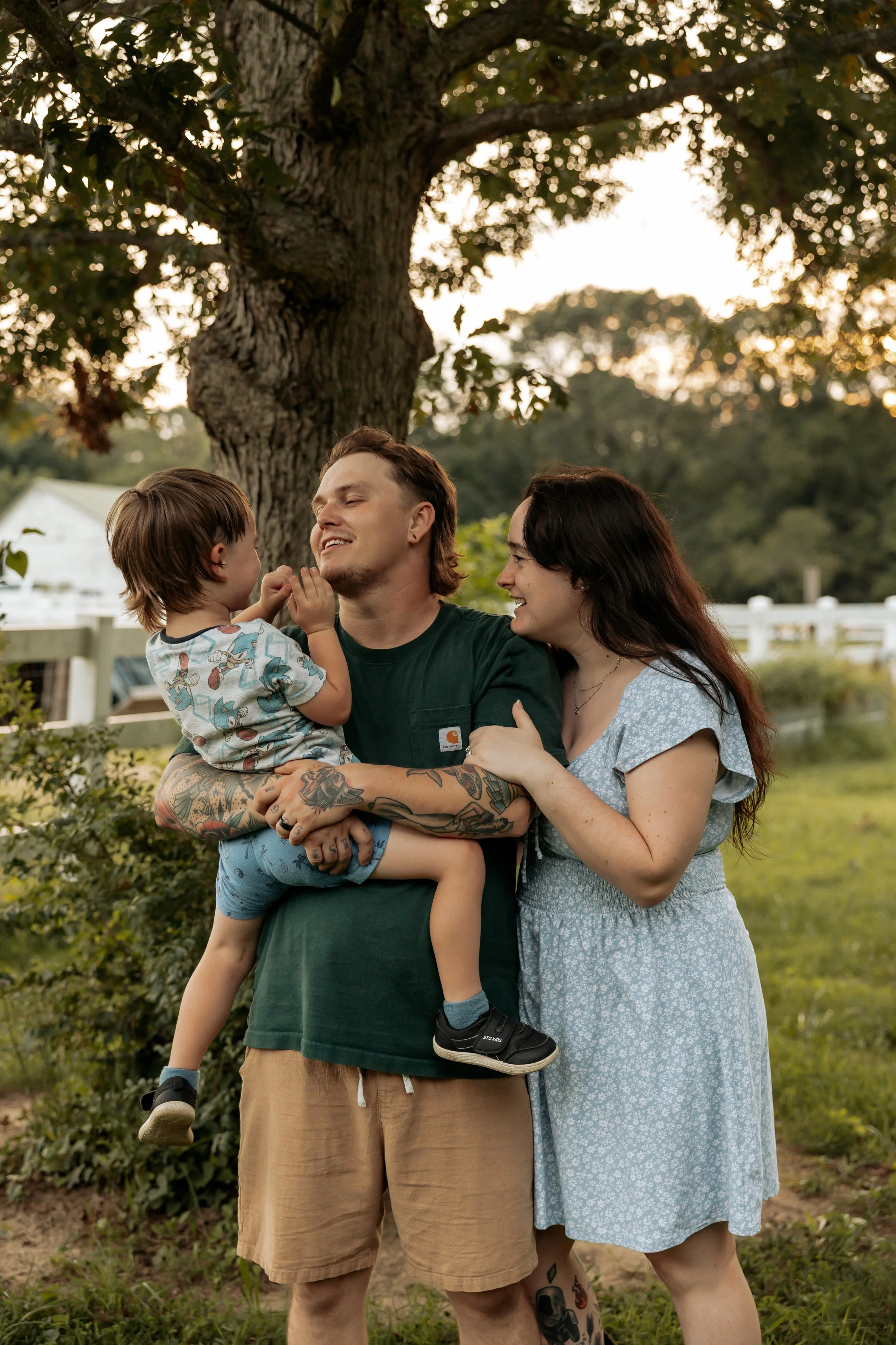 A family of three, consisting of a man with tattoos, a woman, and a young boy, stands outdoors under a large tree during sunset. The man is holding the boy, and they are smiling and interacting affectionately.