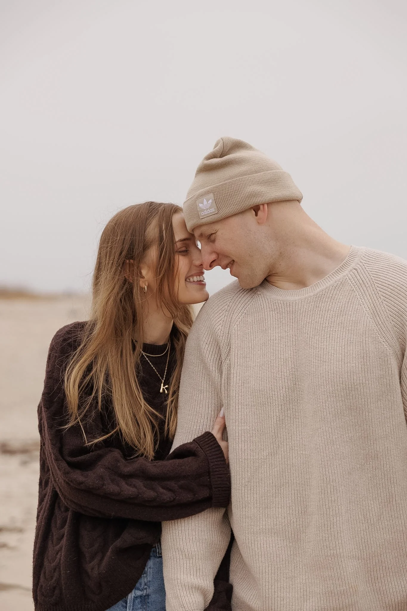 A couple faces touching, smiling, outdoors on a cloudy day, with the woman wearing a dark sweater and the man in a beige beanie and sweater.