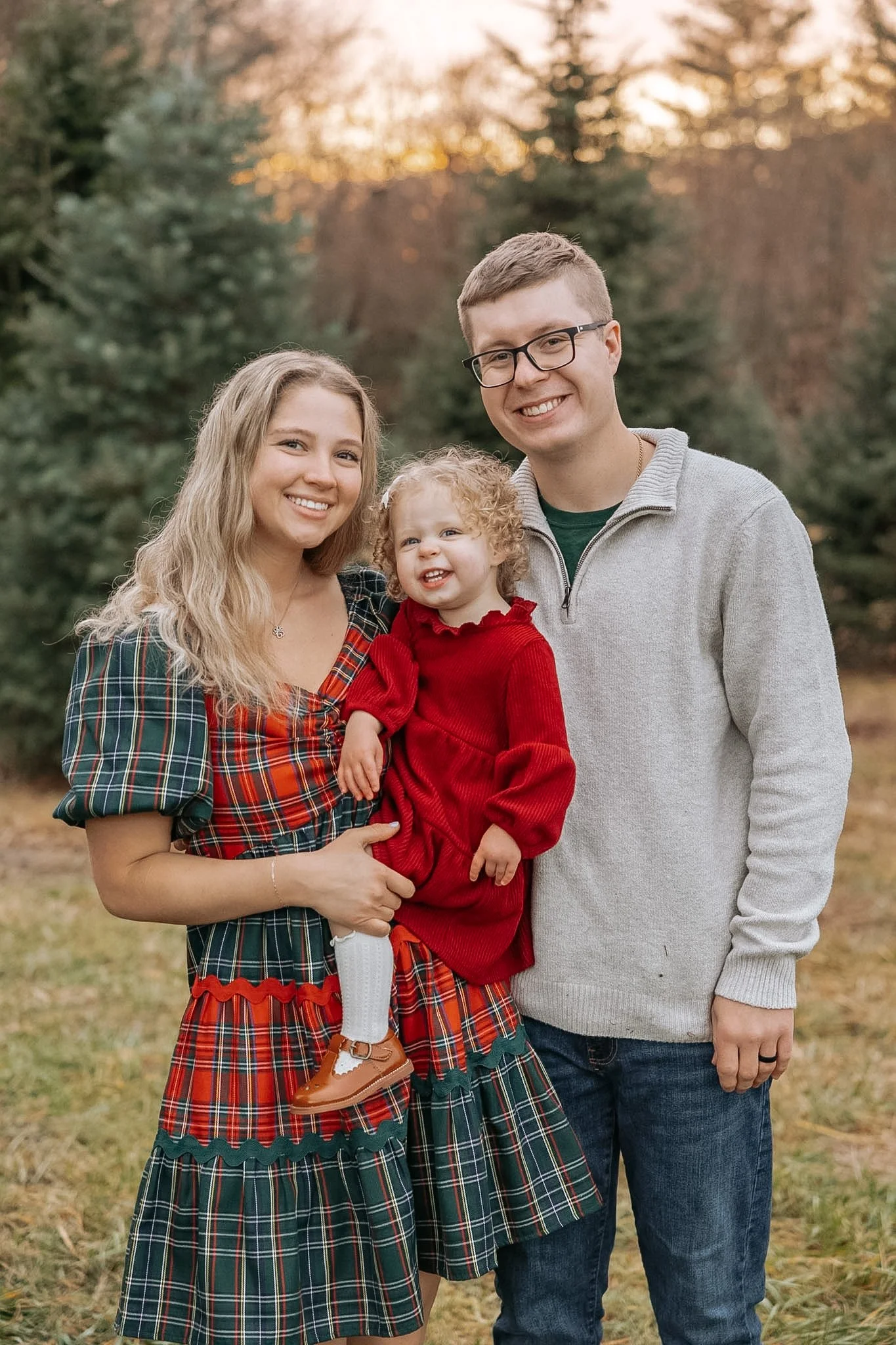 A family of three outdoors during fall, smiling at the camera. The mother has long blonde hair and wears a plaid dress, the father has short hair and wears glasses and a light gray sweater, and their young daughter with curly blonde hair is in a red dress, sitting on her mother’s arm.