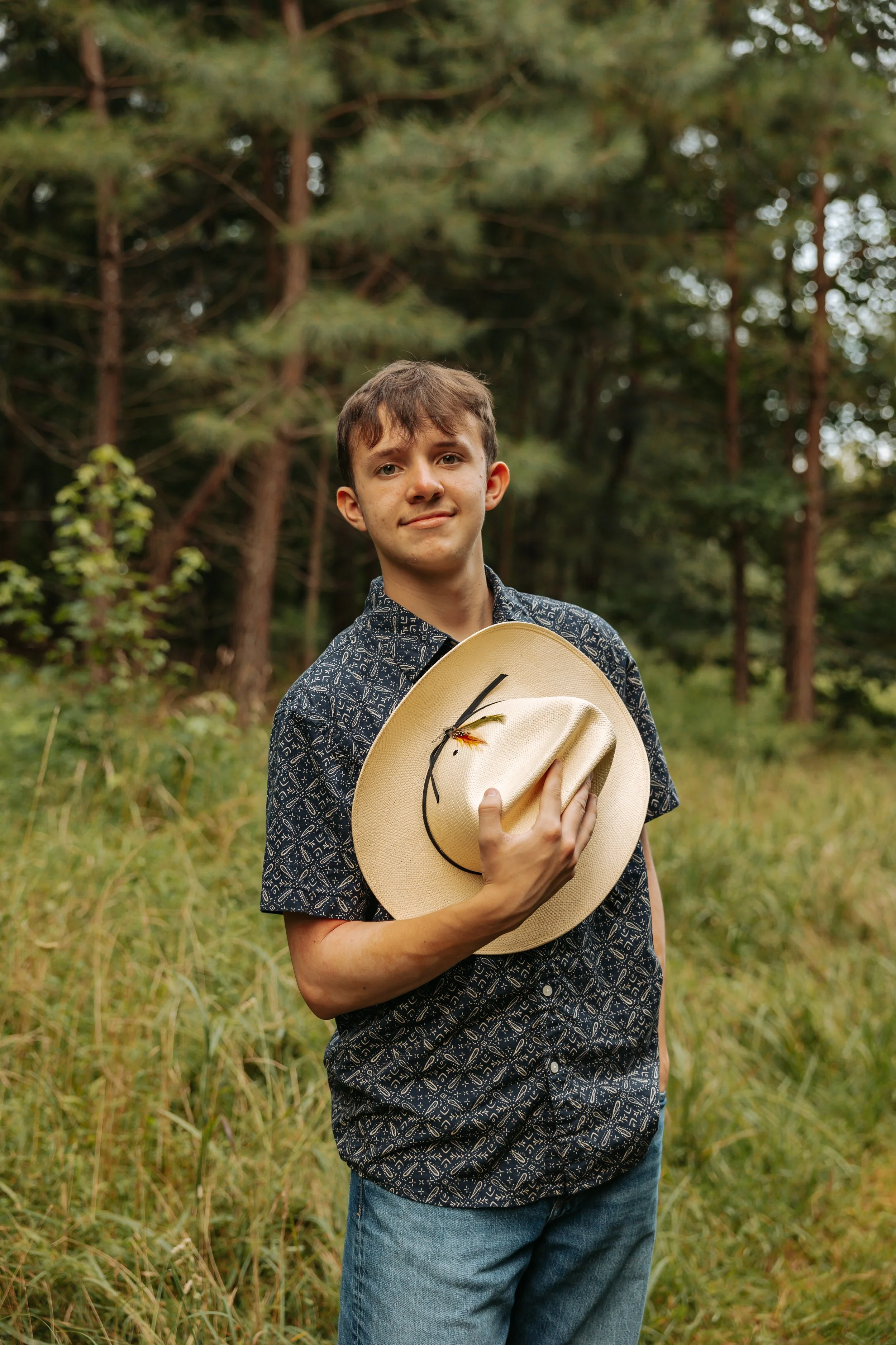 A young man holding a beige hat with a dragonfly on it in a forested area with tall trees and green grass.