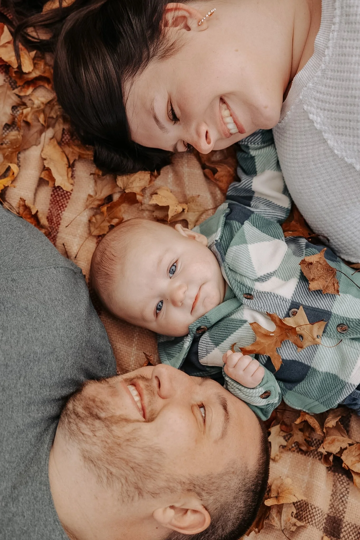 A family of three lying on a blanket covered with autumn leaves, smiling and looking at each other.