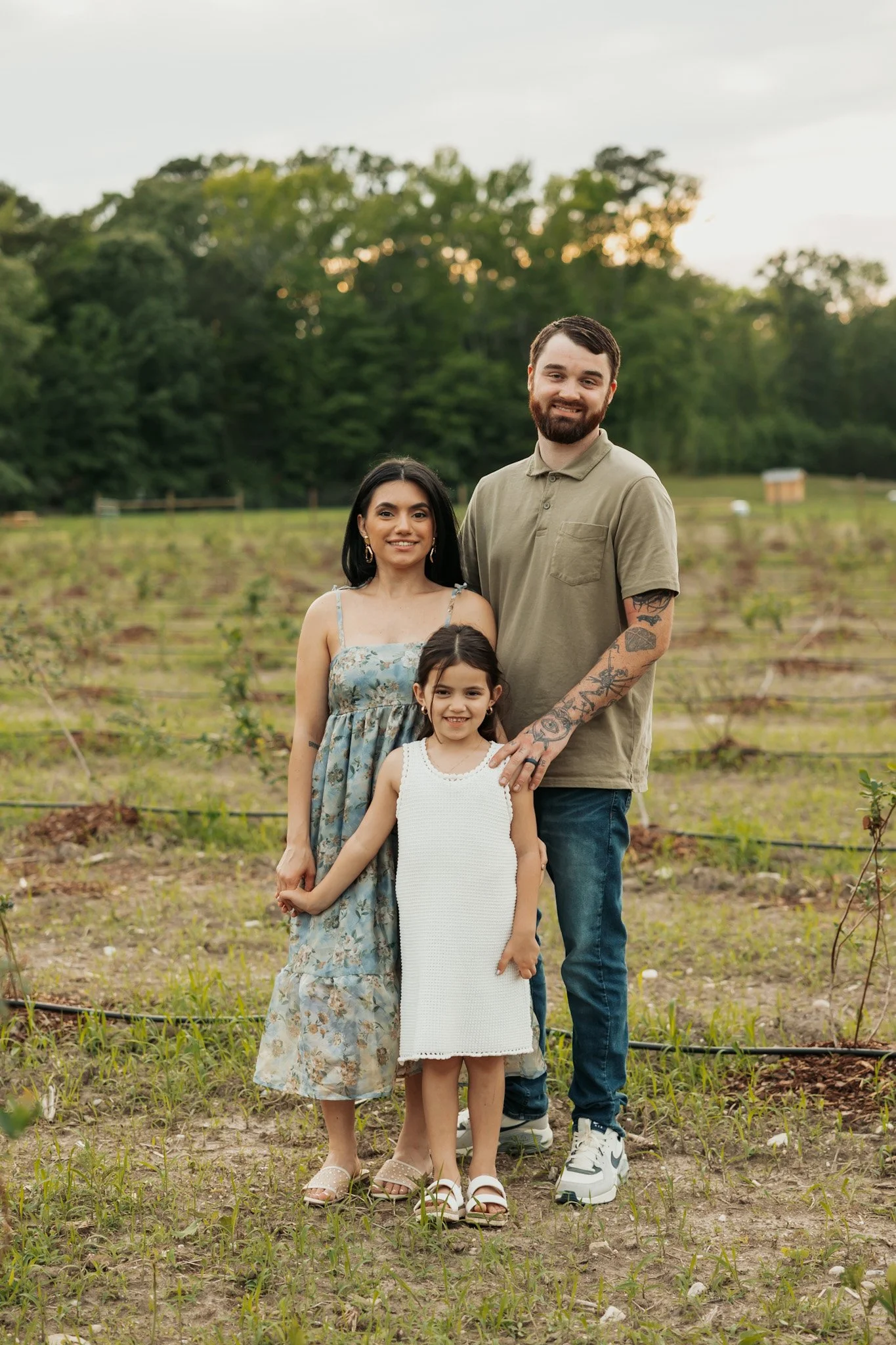 A family of three standing outdoors in a field with green trees in the background. The group includes a man with a beard and tattoos, a woman with long dark hair, and a young girl with dark hair, all smiling at the camera.