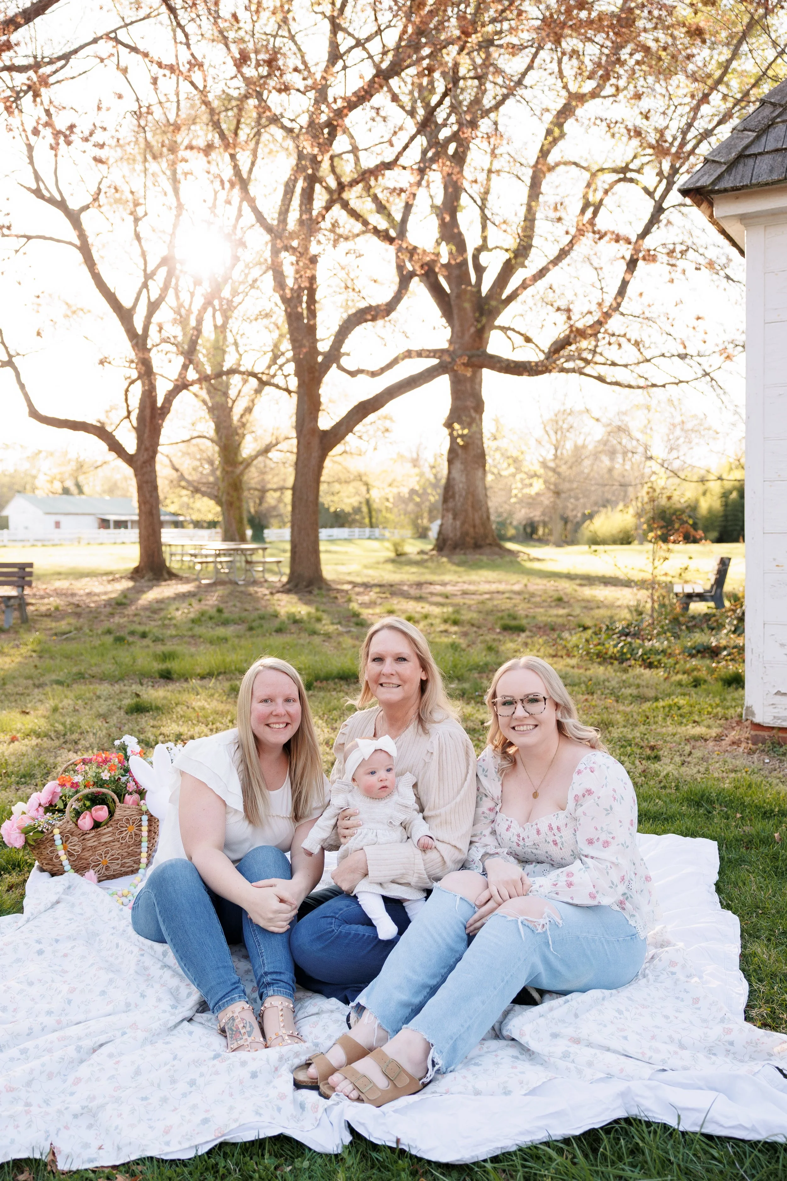 Four women, one of whom is a baby, sitting outdoors on a white blanket during golden hour with trees and a house in the background, enjoying a sunny day.