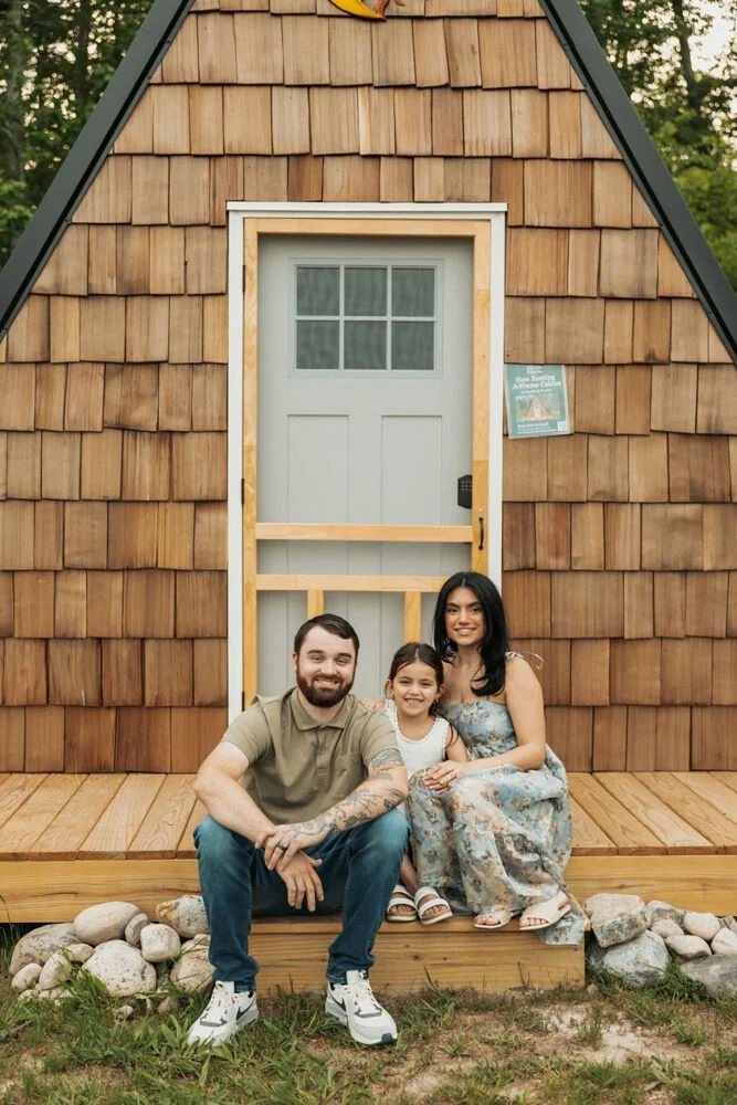 A family of three sitting on a wooden porch in front of a tiny wooden house with a small screen door. The family includes a man, woman, and young girl, smiling at the camera.