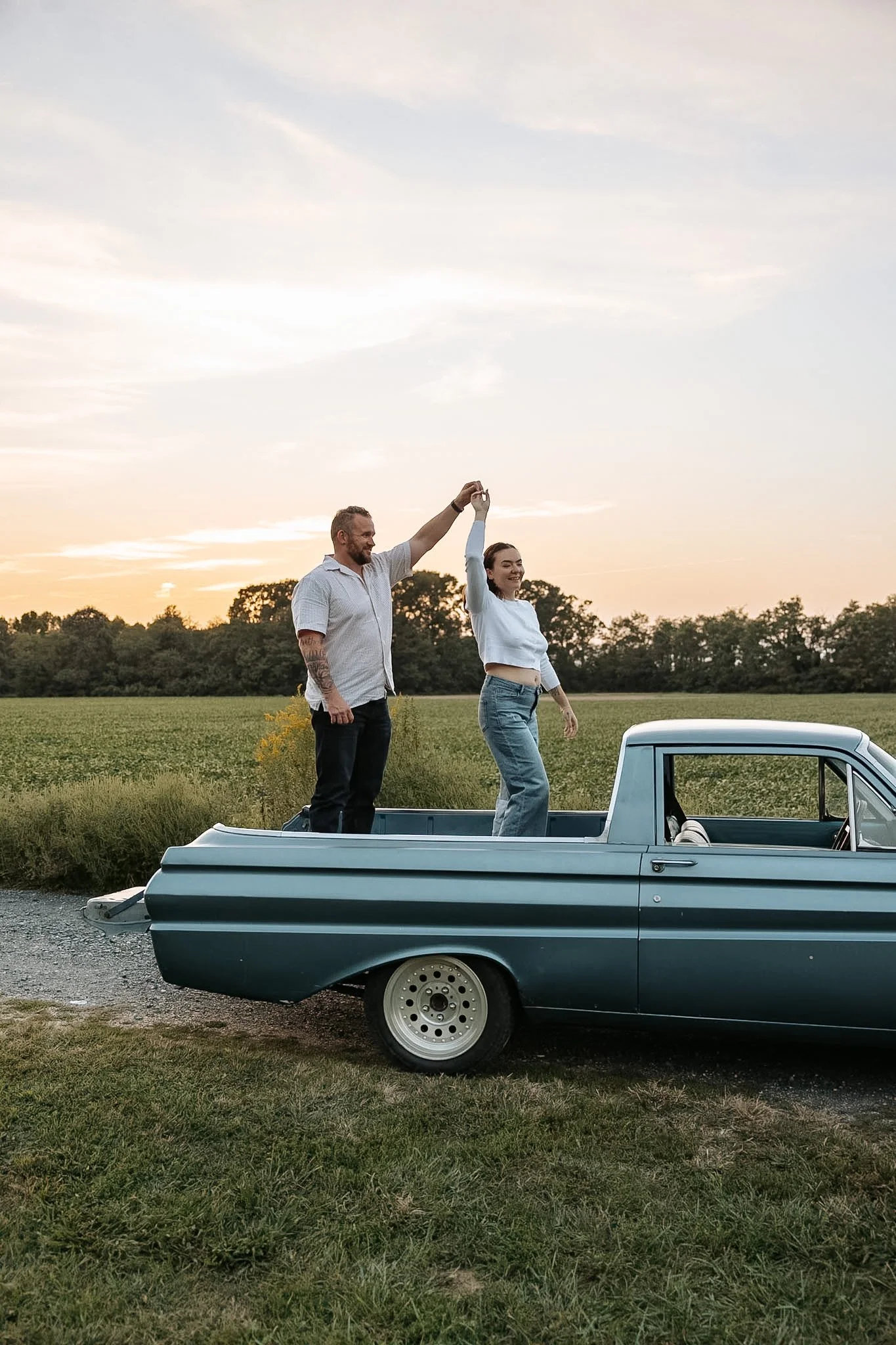 A man and woman standing in the bed of a vintage pickup truck, dancing and holding hands with the sunset in the background.