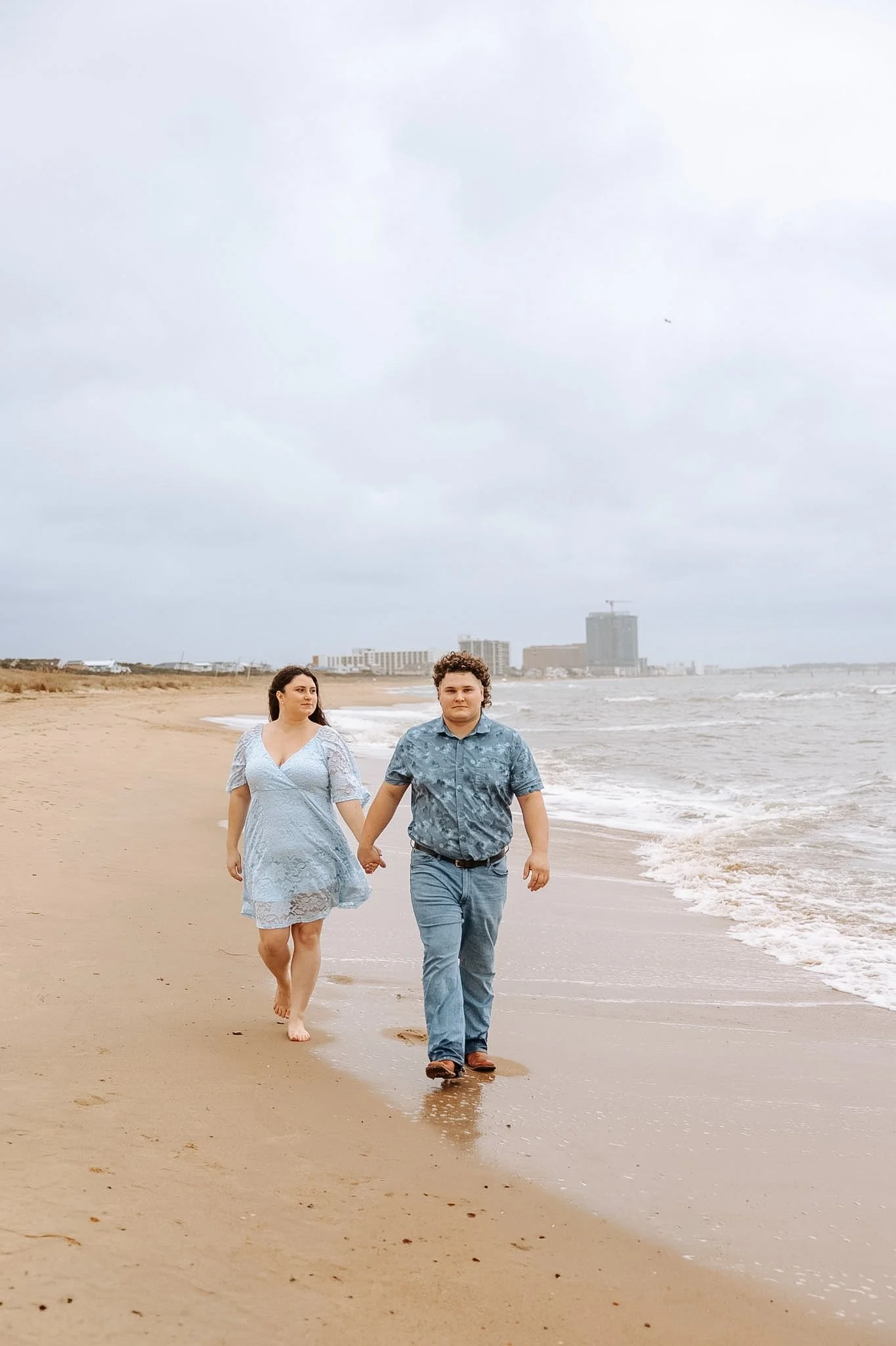 A couple walking hand in hand on a sandy beach during cloudy weather, with city buildings in the background.
