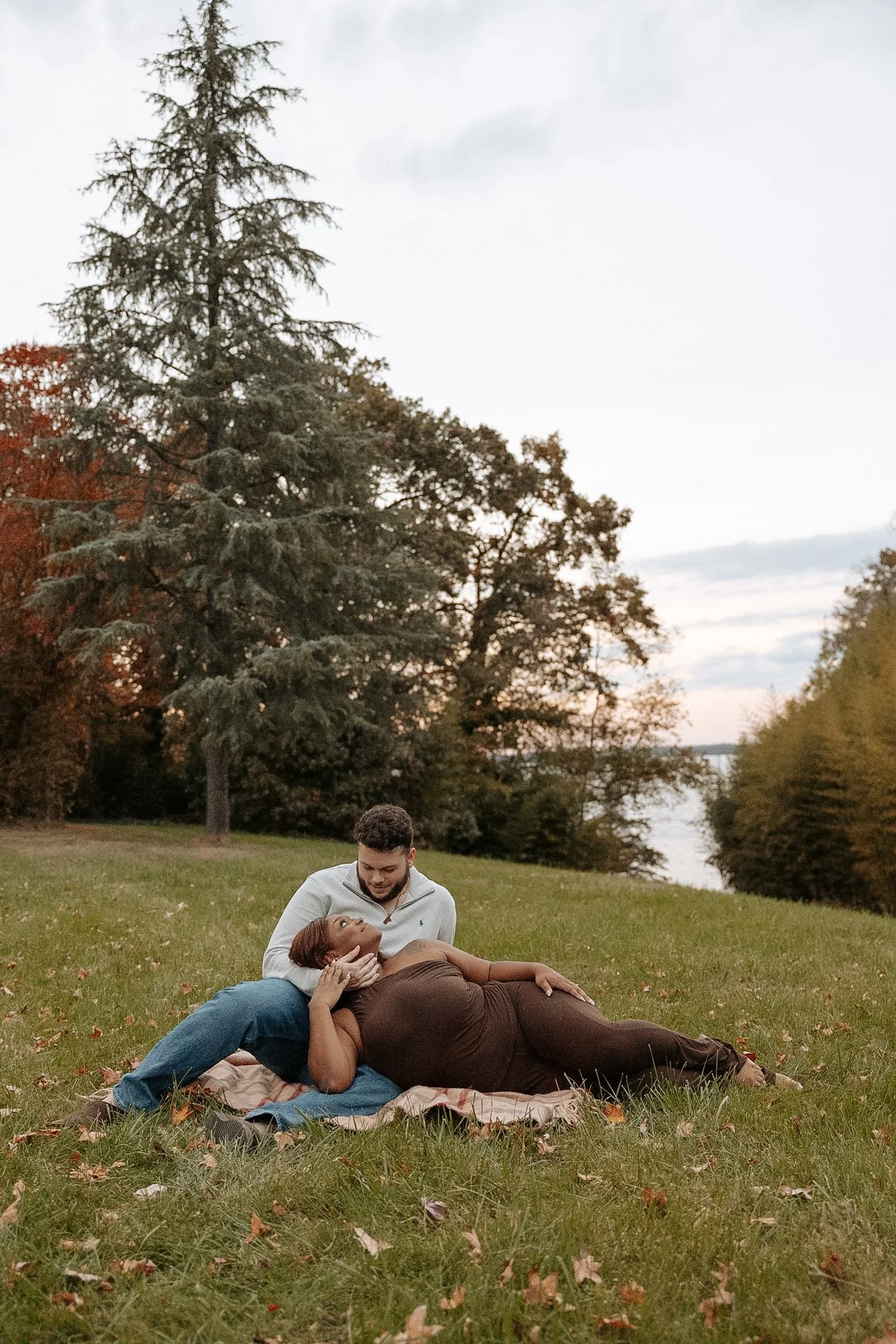 A couple sitting on a blanket on the grass in a park near a river, with trees in the background, during fall. The woman, is reclining and looking at the man, who is sitting beside her. They appear happy and close.
