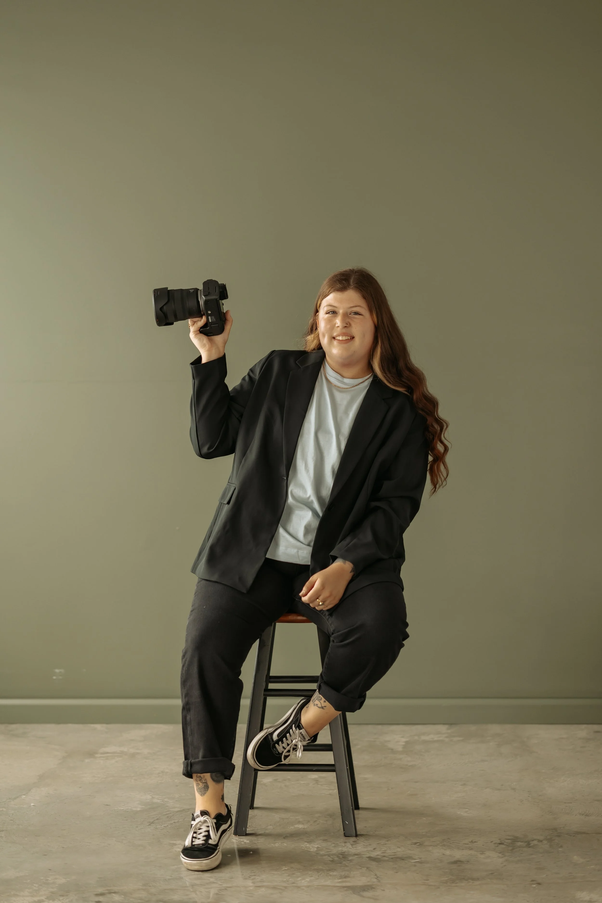 A woman with long, wavy brown hair, wearing a black blazer, blue t-shirt, black jeans, and black Vans sneakers, sitting on a stool and holding a camera in her right hand against a plain green background.