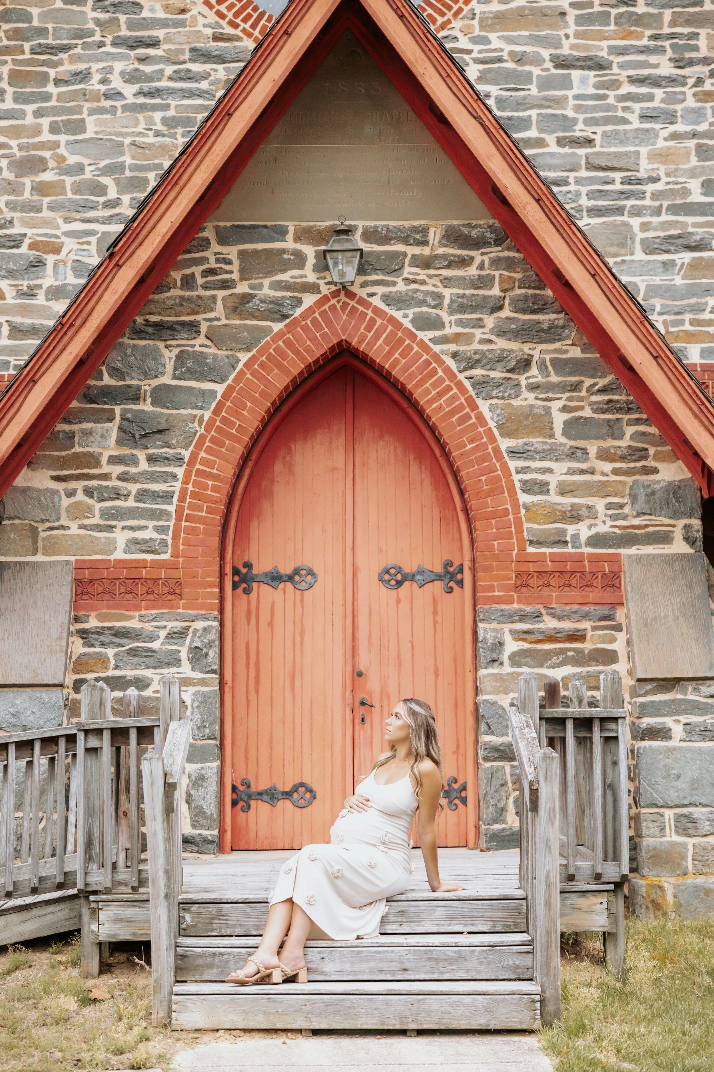 A pregnant woman in a white dress sitting on wooden steps outside a stone church with large red door and brick arch, looking to the side.