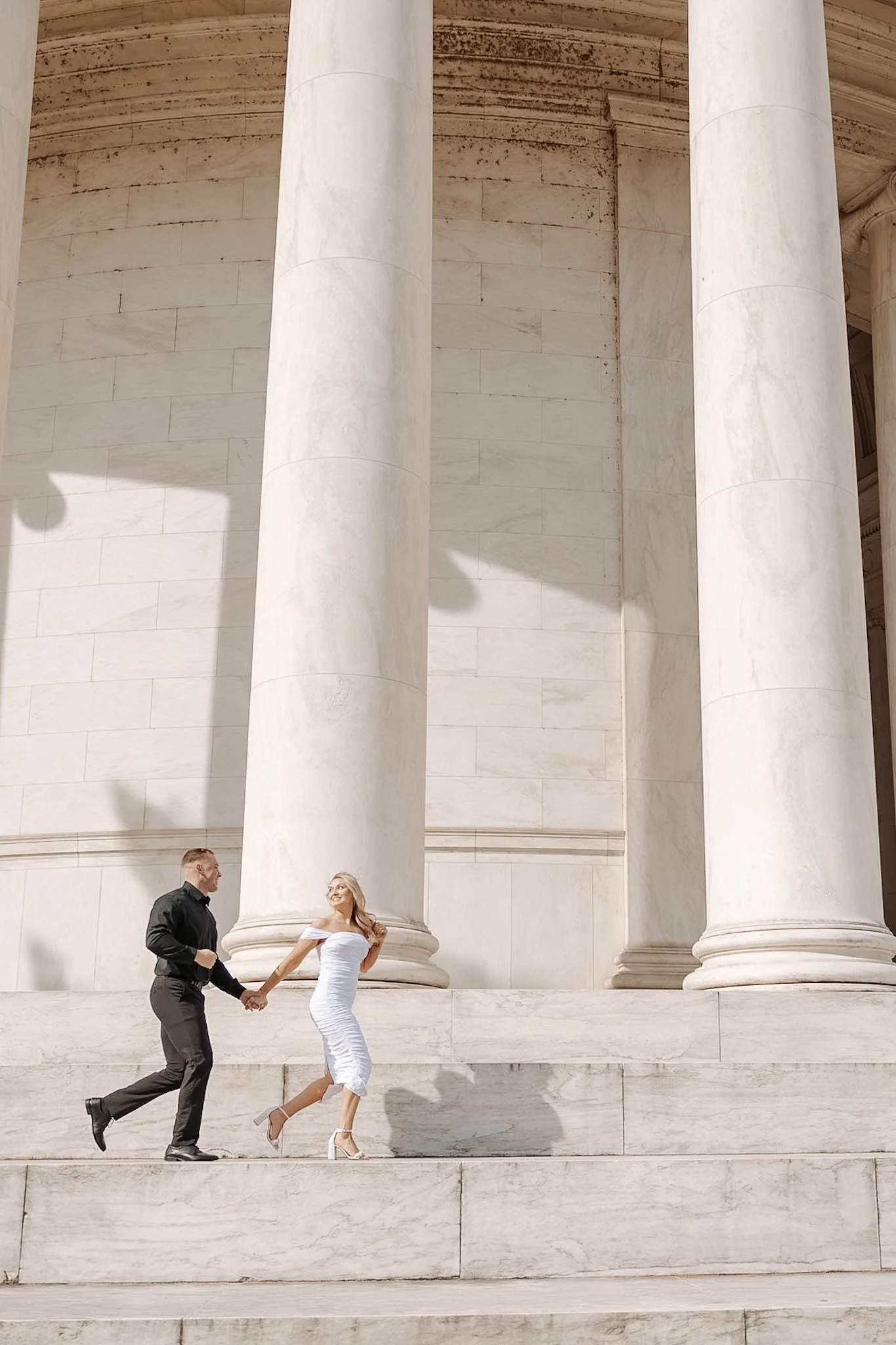 A man and woman holding hands and running up marble steps in front of large marble columns at the Lincoln Memorial.