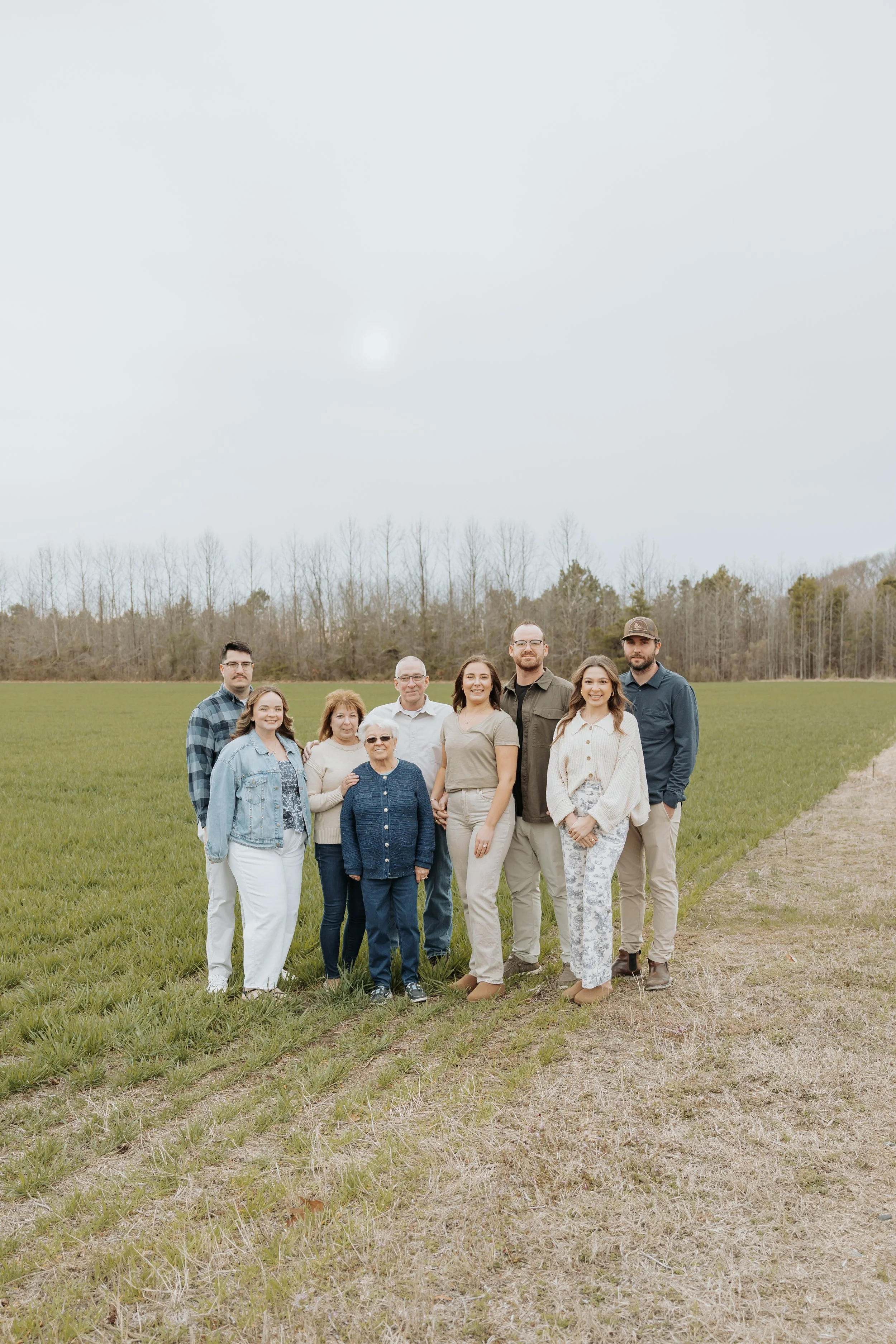 A multi-generational family standing outdoors on a grassy field, with trees in the background and overcast sky.