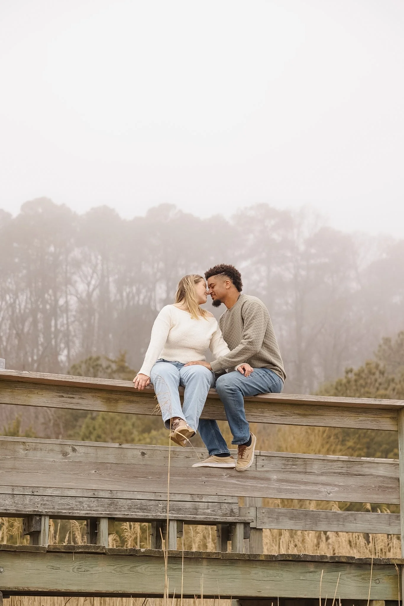 A couple sitting on a wooden railing outdoors, about to kiss, with a foggy background and trees.