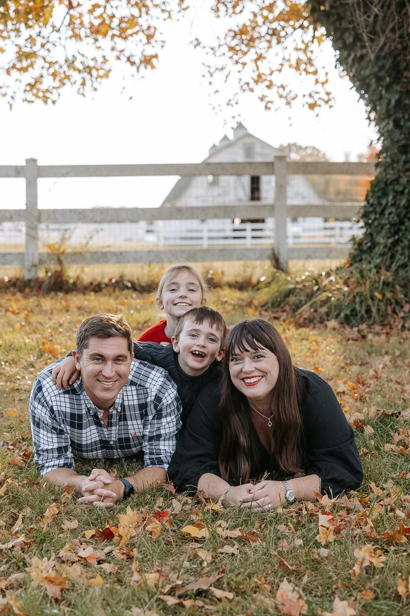 Family of four lying on grass covered with autumn leaves, smiling, with a white fence and barn in the background.