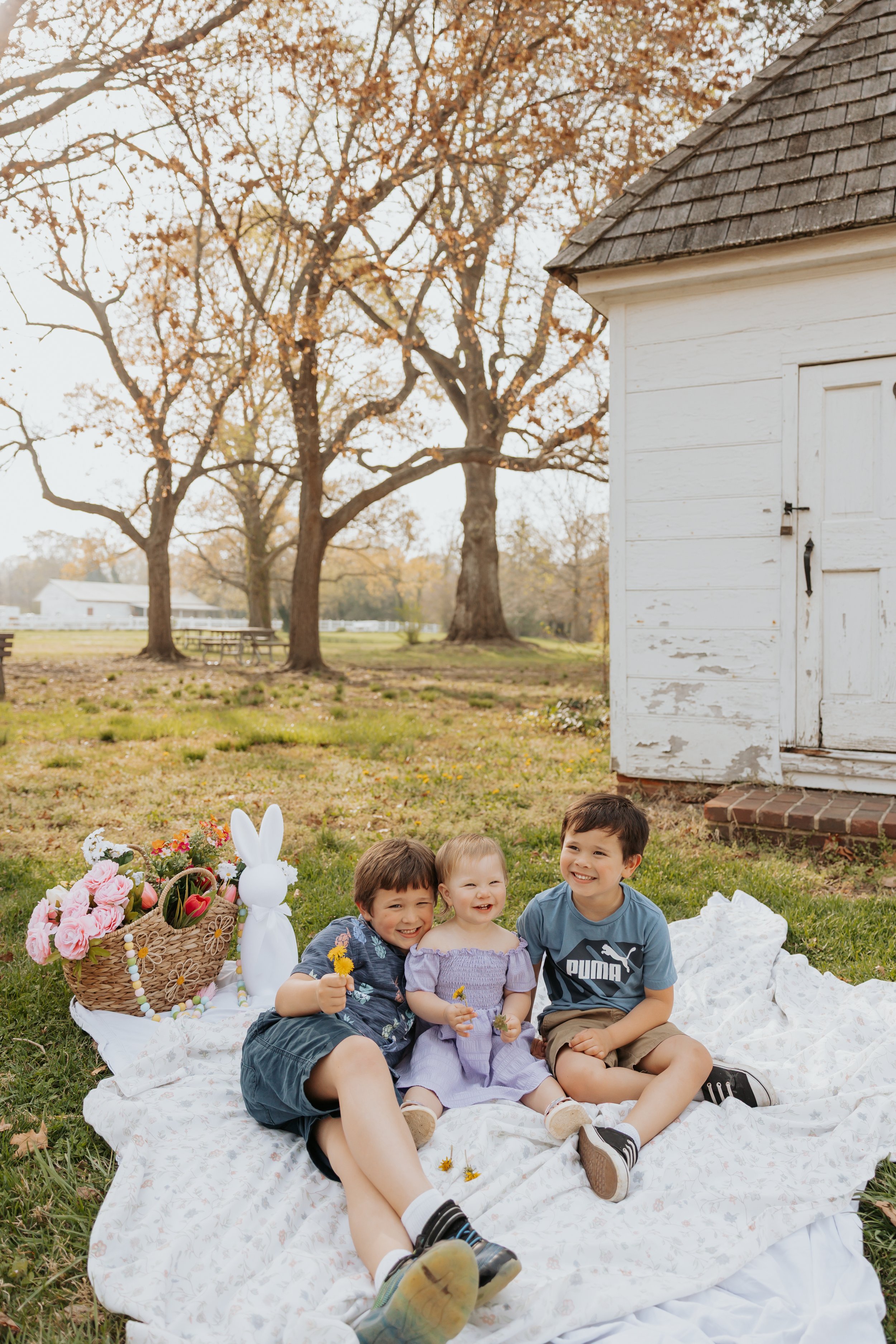Three children sitting on a white blanket outdoors during daytime with a basket of flowers and decorated bunny figure, trees and a white wooden shed in the background.