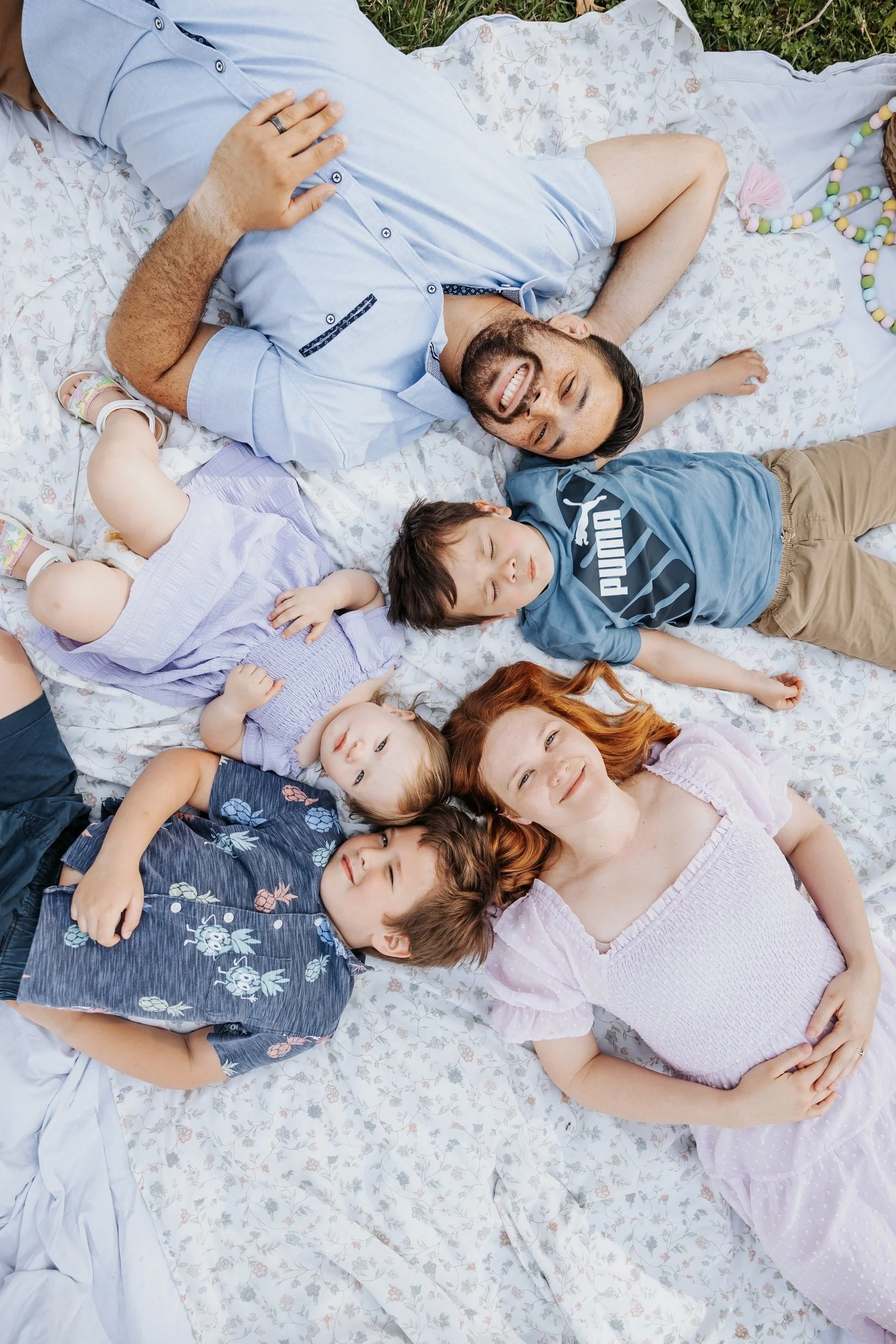 A family lying on a floral-patterned blanket outdoors, smiling and relaxing
