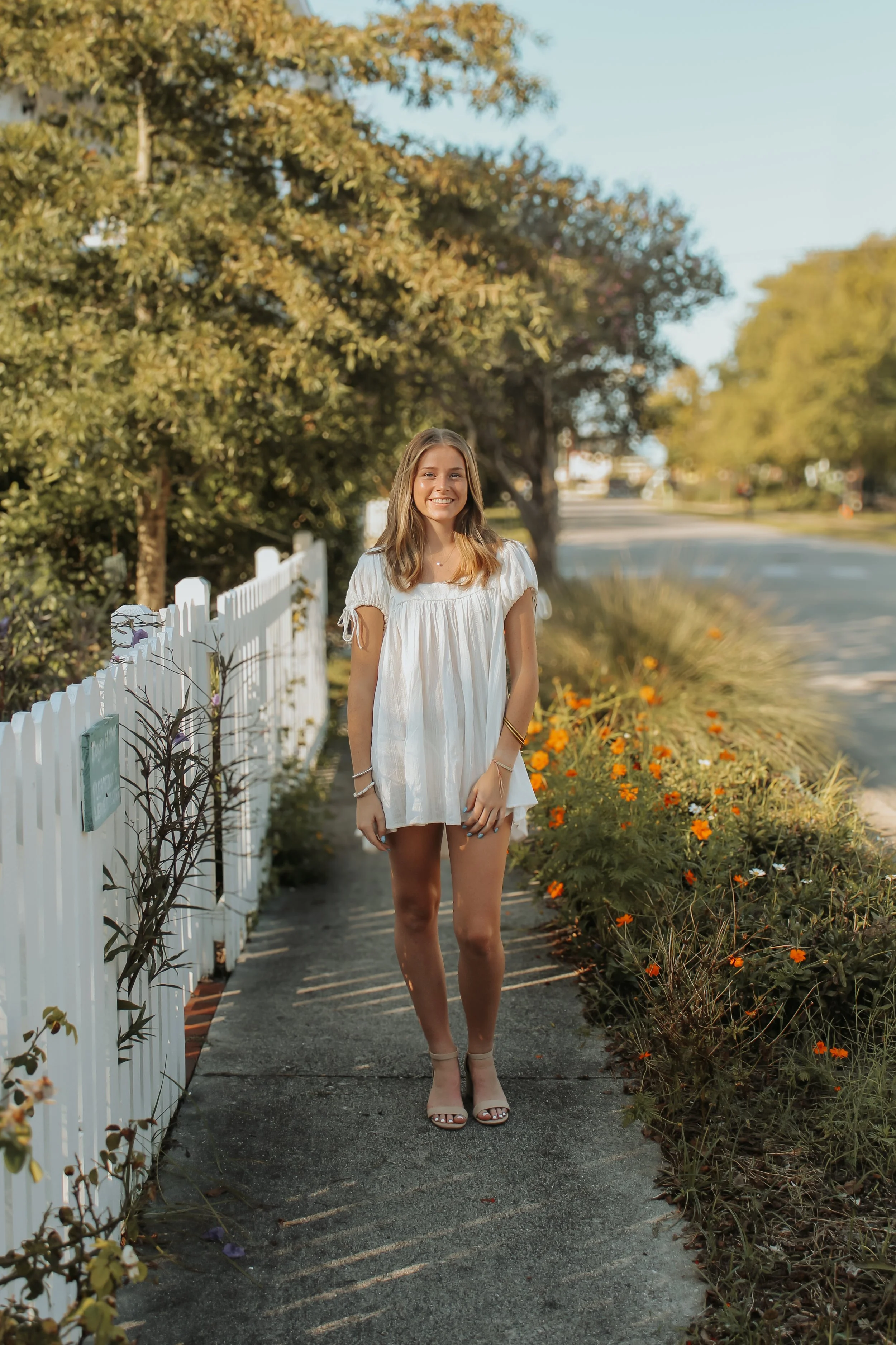 A young woman smiling stands on a sidewalk next to a white picket fence and orange flowers, with trees and a street in the background.