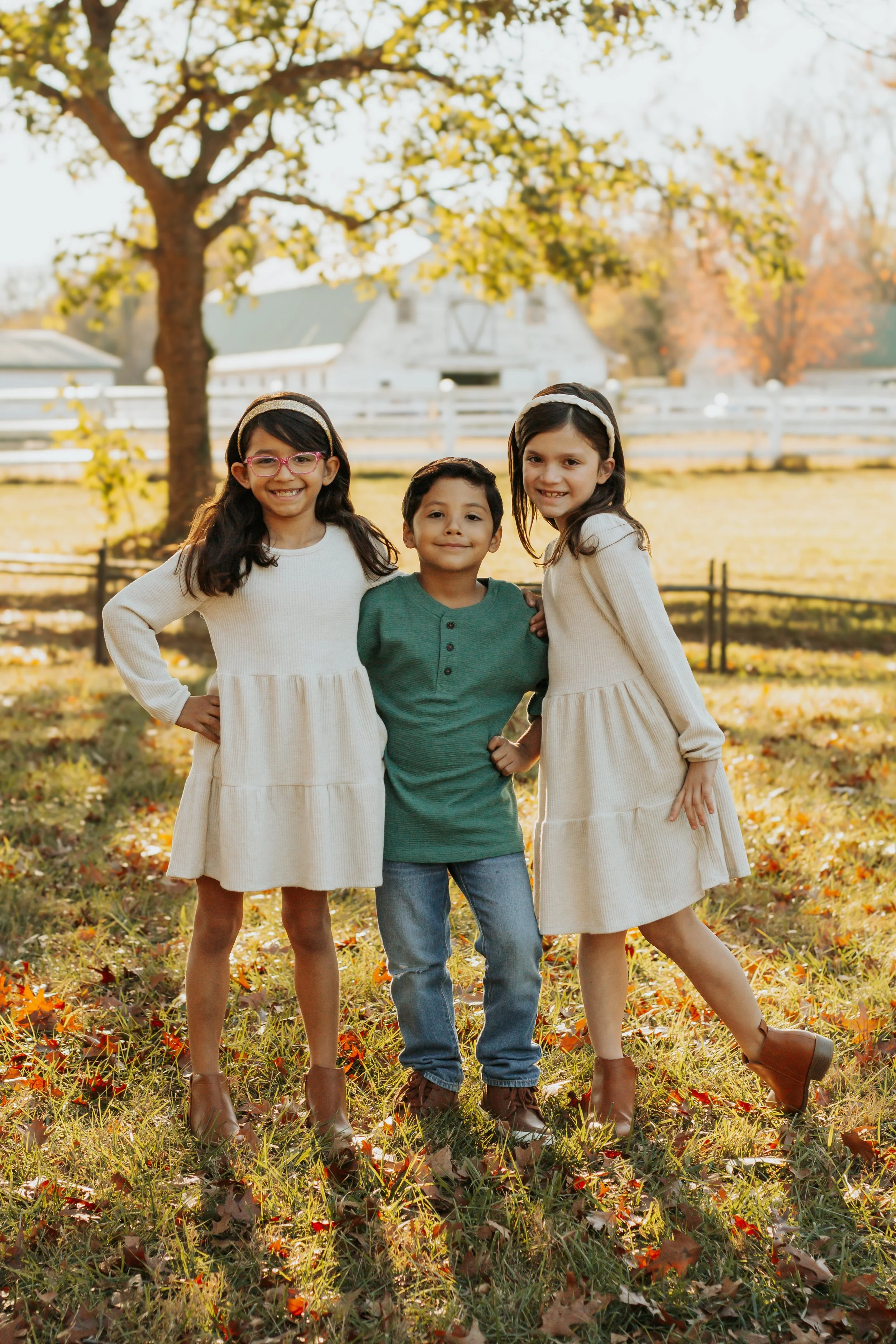 Three children, two girls and one boy, standing together on a grassy fall day in front of a tree, with a white fence and a house in the background.
