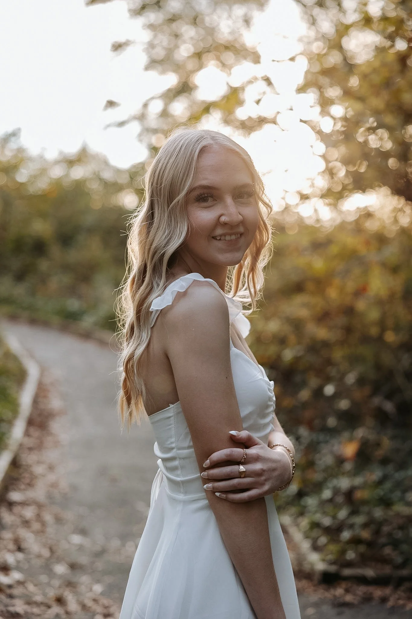 A young woman with blonde hair in a white dress standing outdoors on a trail with trees in the background, backlit by the setting sun.