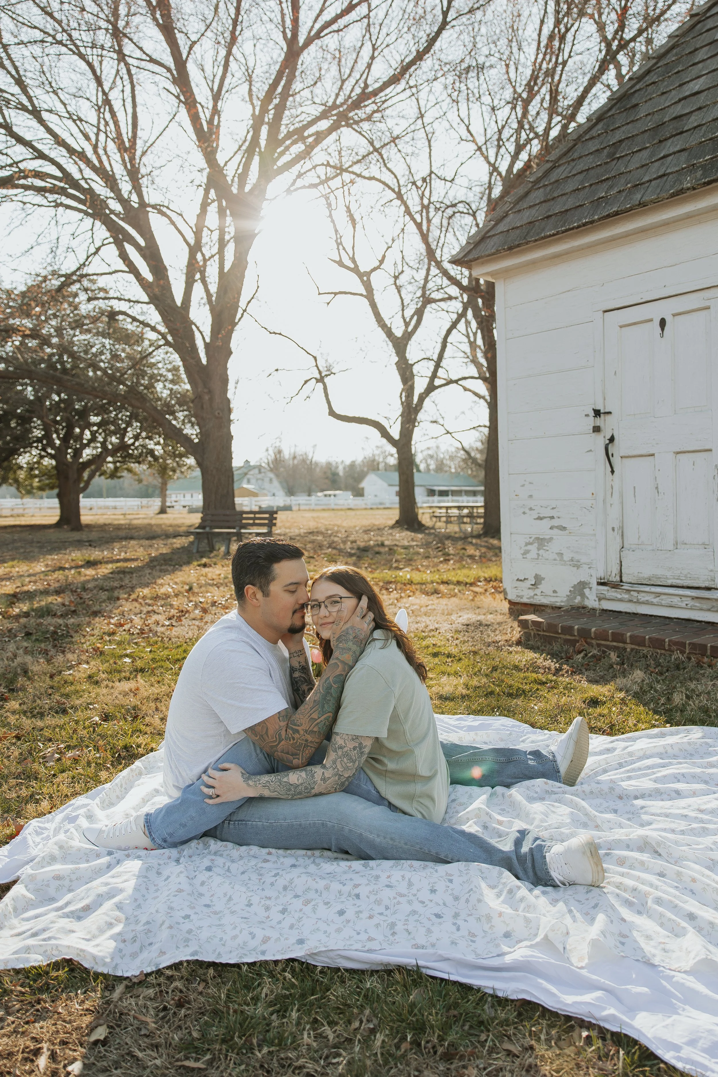 A couple sitting on a blanket outdoors near a white shed, with leafless trees and sunlight in the background.