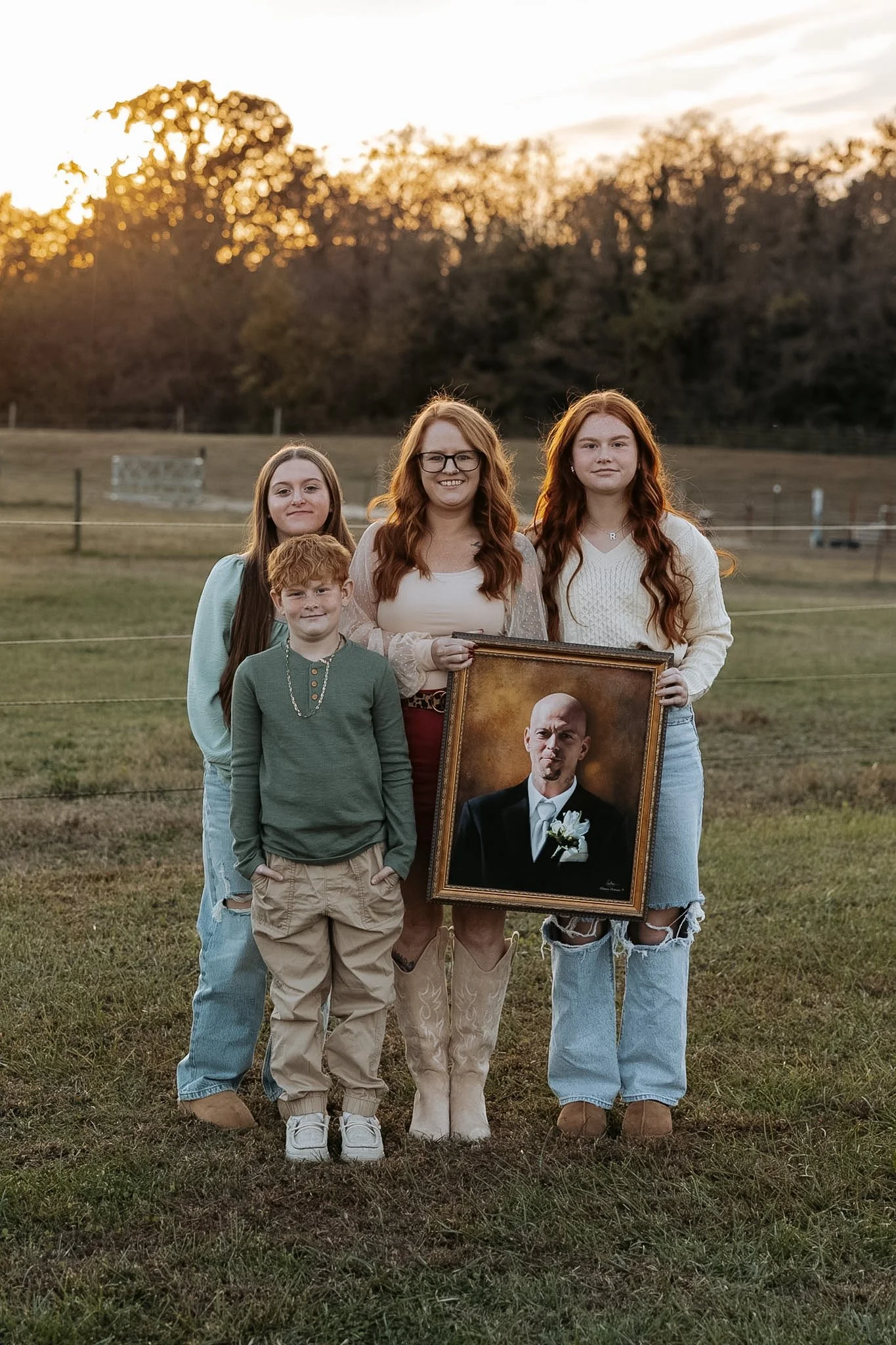 Group of five people, three women and two children, standing outdoors on grass during sunset, holding a framed portrait of a man in a suit with a white flower boutonniere.