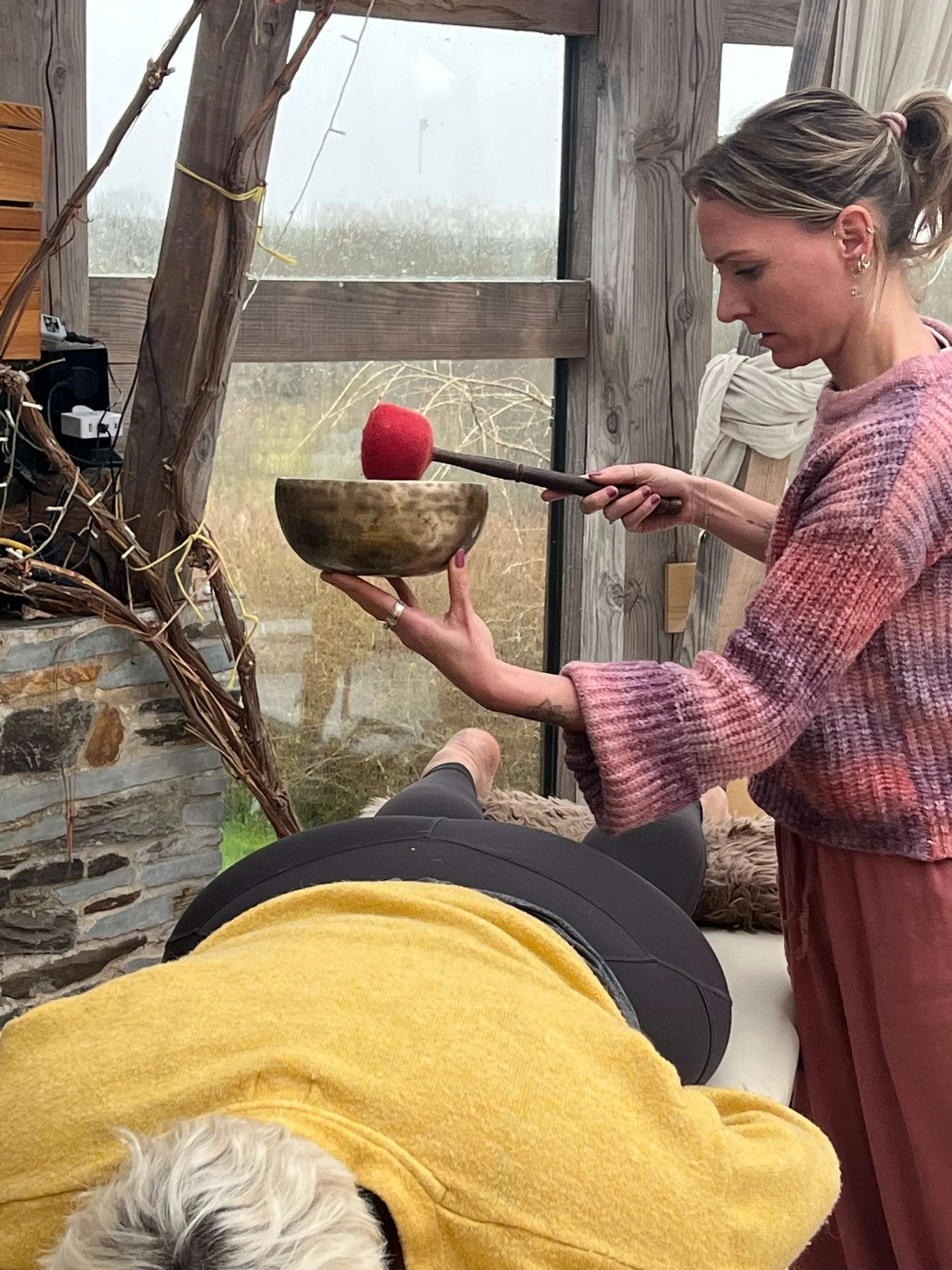 A woman in a plaid shirt holds a wooden bowl and a mallet with a felted red ball on top, next to a person lying on a bench wearing a yellow sweater, with a dog partially visible in the foreground.