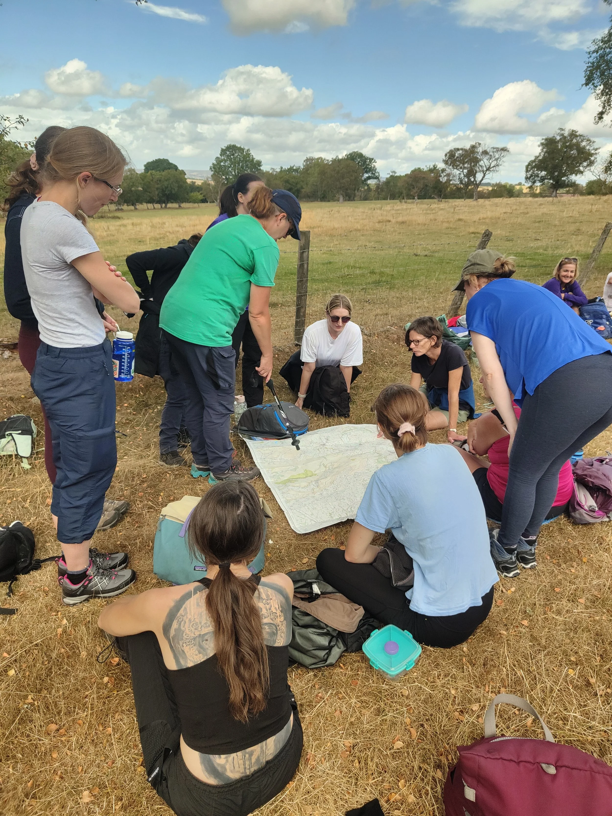 A group of people gathered around a map on the ground in an open field with trees and a cloudy sky in the background.