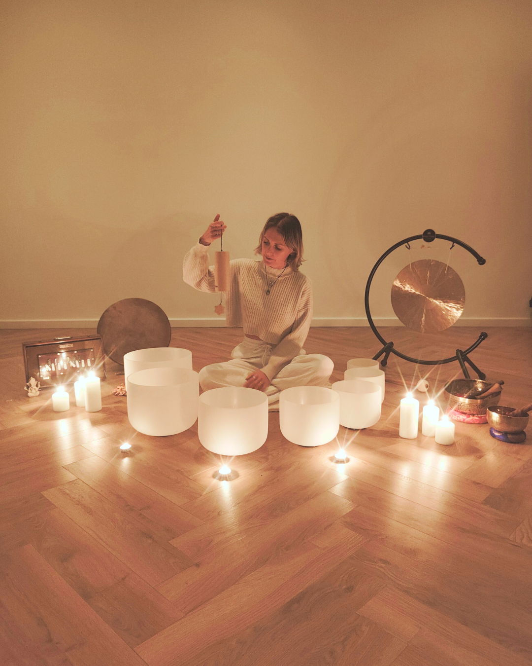 A woman in a white sweater sitting on a wooden floor surrounded by candles and singing bowls, with a large gong and a frame with candles beside her, in a serene setting.