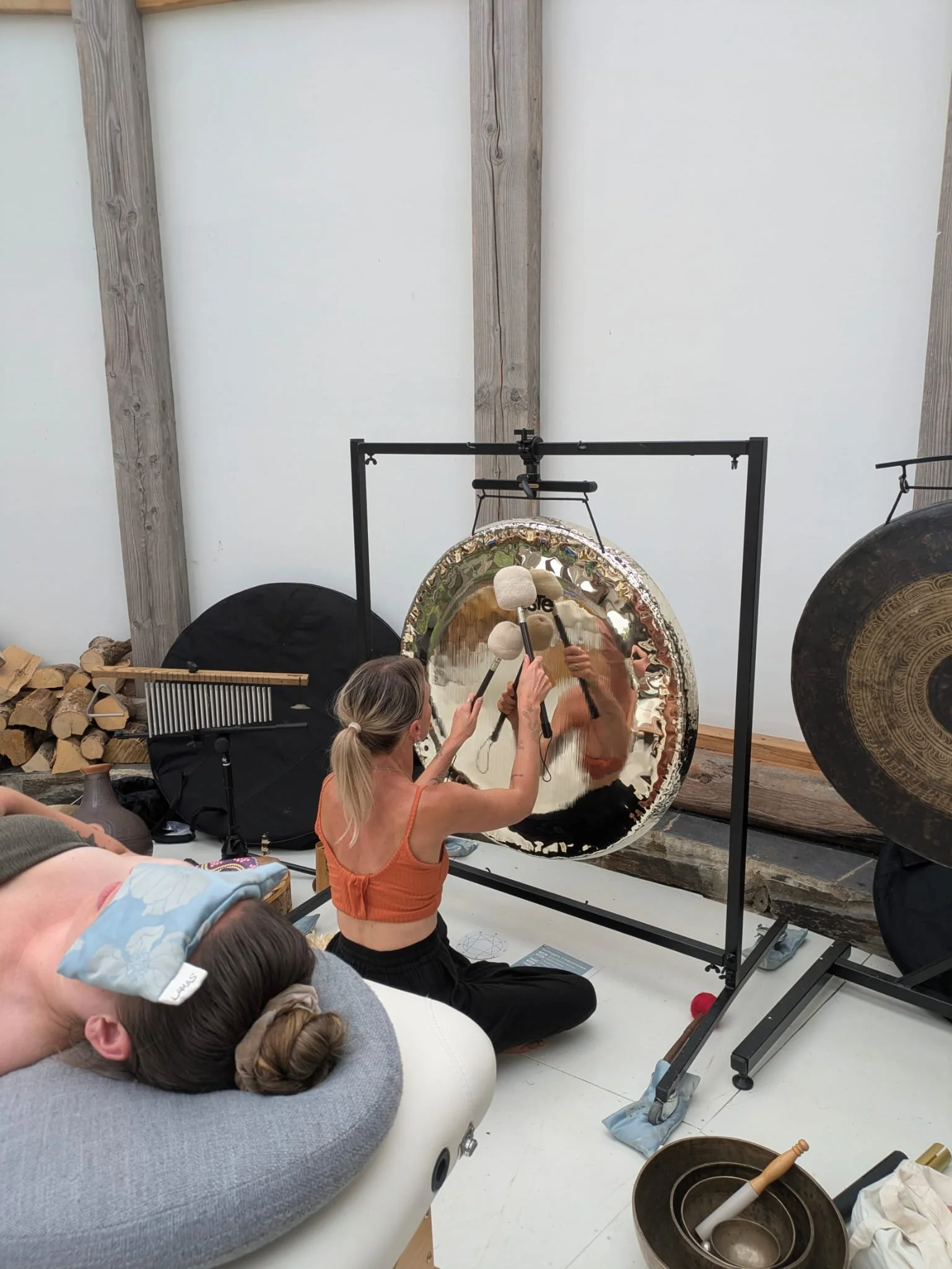 A woman playing a large gong while a person is resting nearby with a cloth over their face, in an indoor setting.