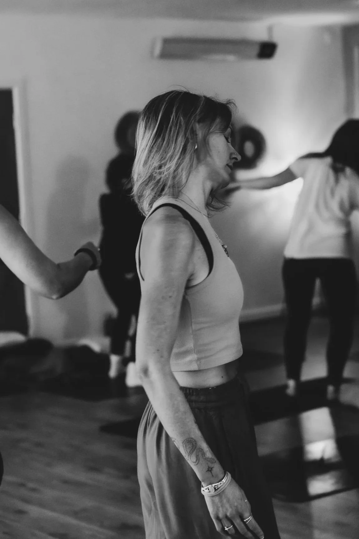 A woman with shoulder-length hair and tattoos on her arm practicing yoga in a studio, with other people in the background.