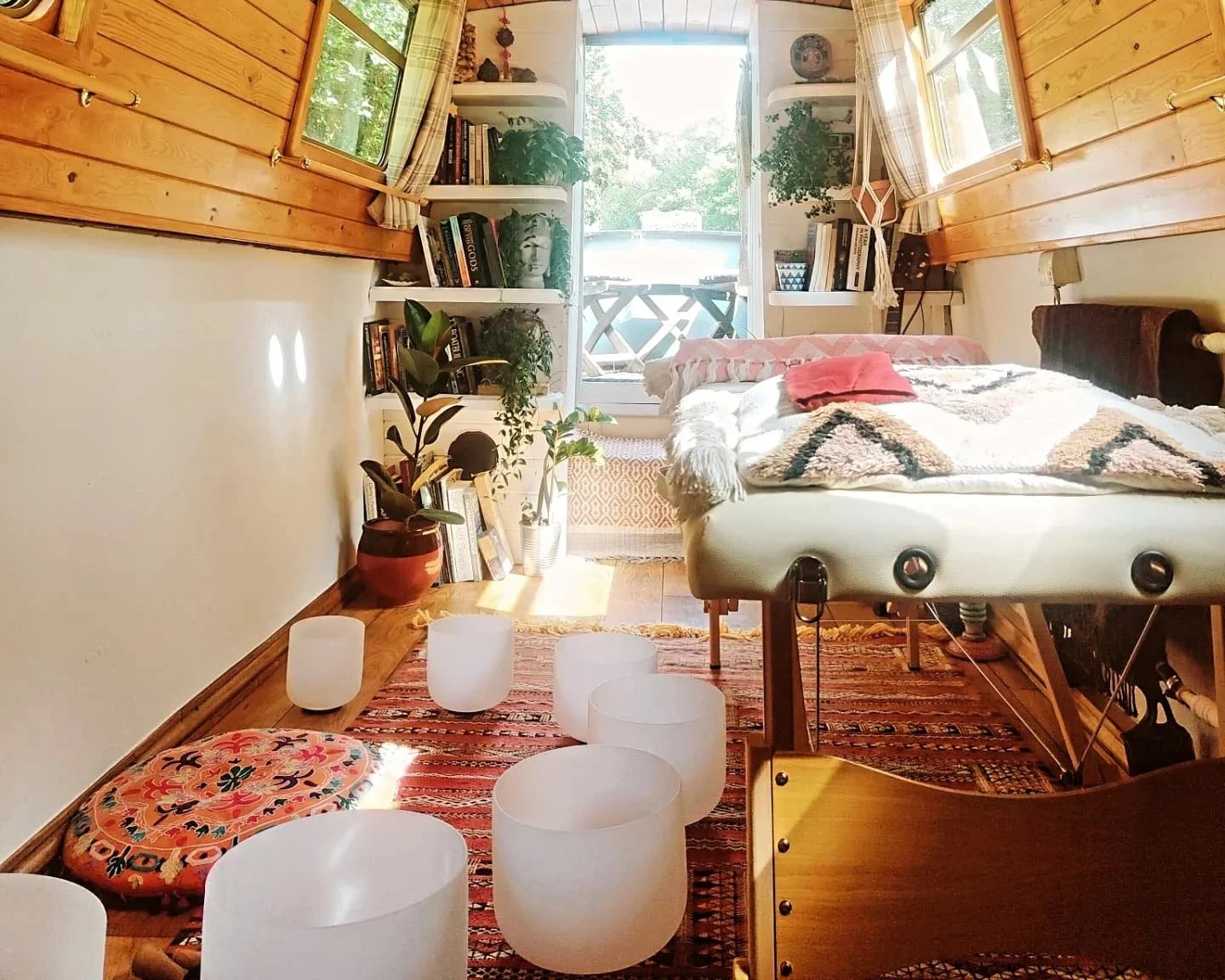 Bright bedroom with wooden walls, a bed with a blanket and pillow, bookshelves, potted plants, and a balcony with a view outside.