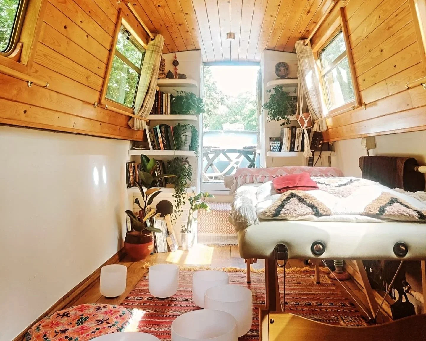A cozy bedroom with wood-paneled walls and ceiling, a bed with a blanket and pillows, potted plants, bookshelves, windows with checkered curtains, and a balcony outside.