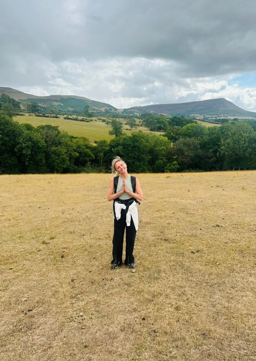 A woman standing in a field with mountains in the background, smiling with hands pressed together in a prayer-like gesture. She is wearing a backpack, a sleeveless top, and black pants.