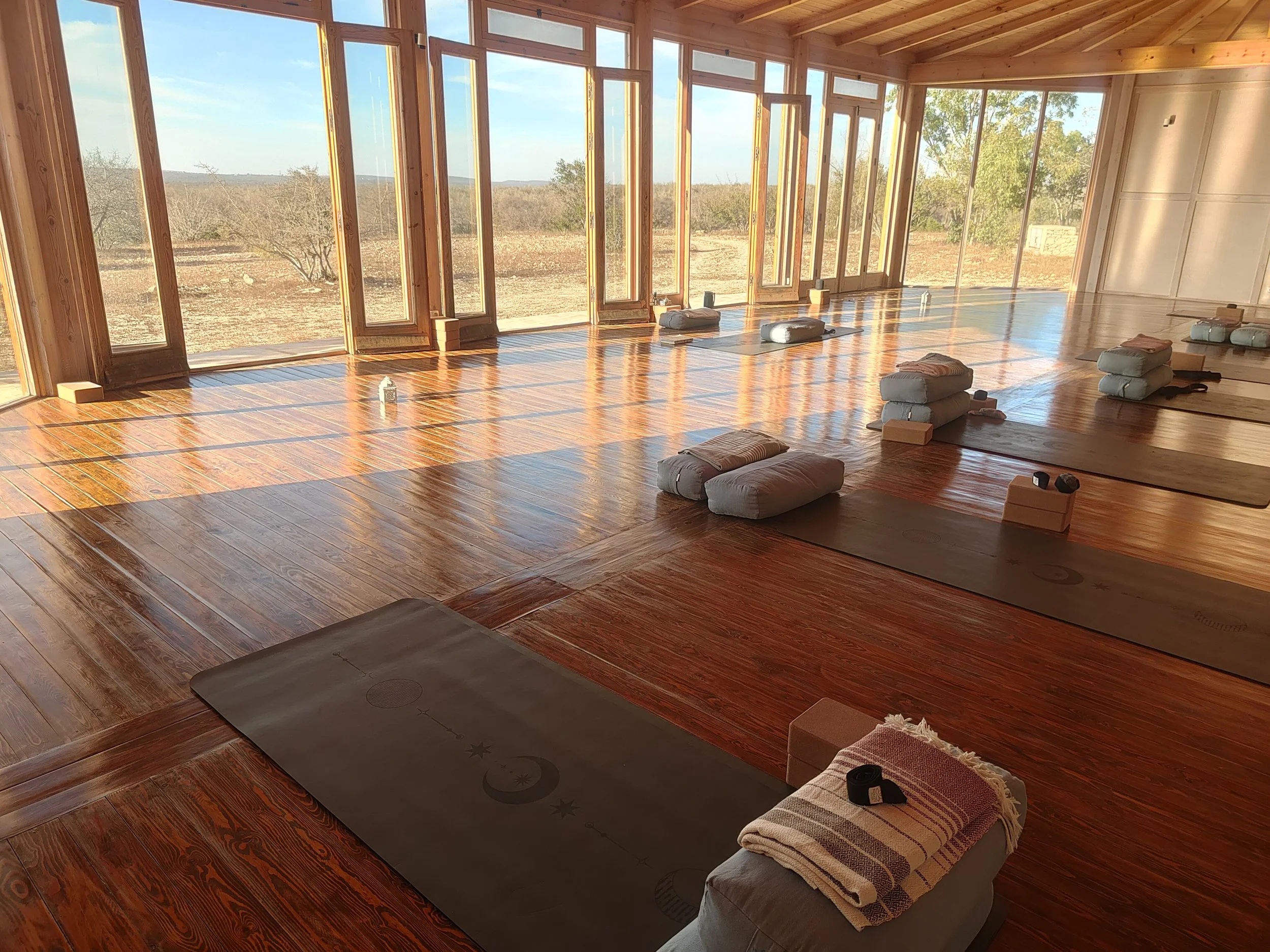 Spacious yoga studio with wooden floors and large windows, six yoga mats with blankets and blocks, and sunlight streaming in.