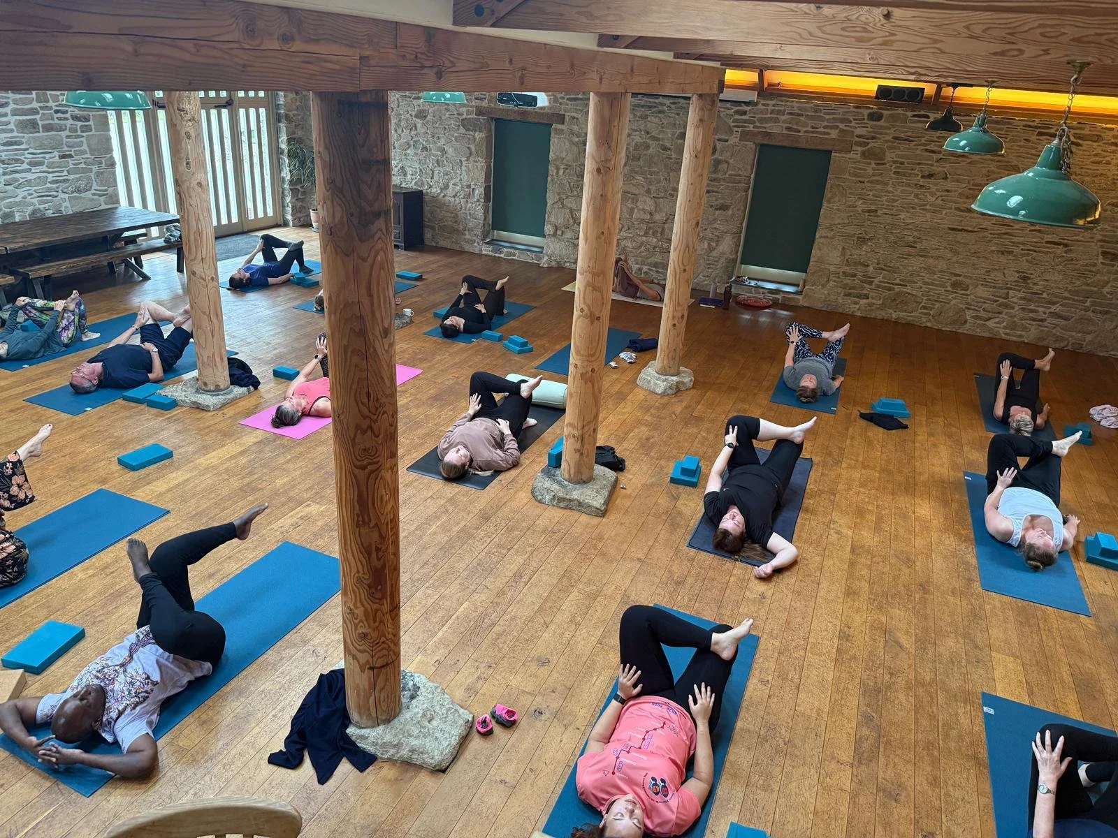 People participating in a yoga class indoors, lying on yoga mats and practicing poses, with some using blocks for support, in a rustic room with wooden beams and stone walls.