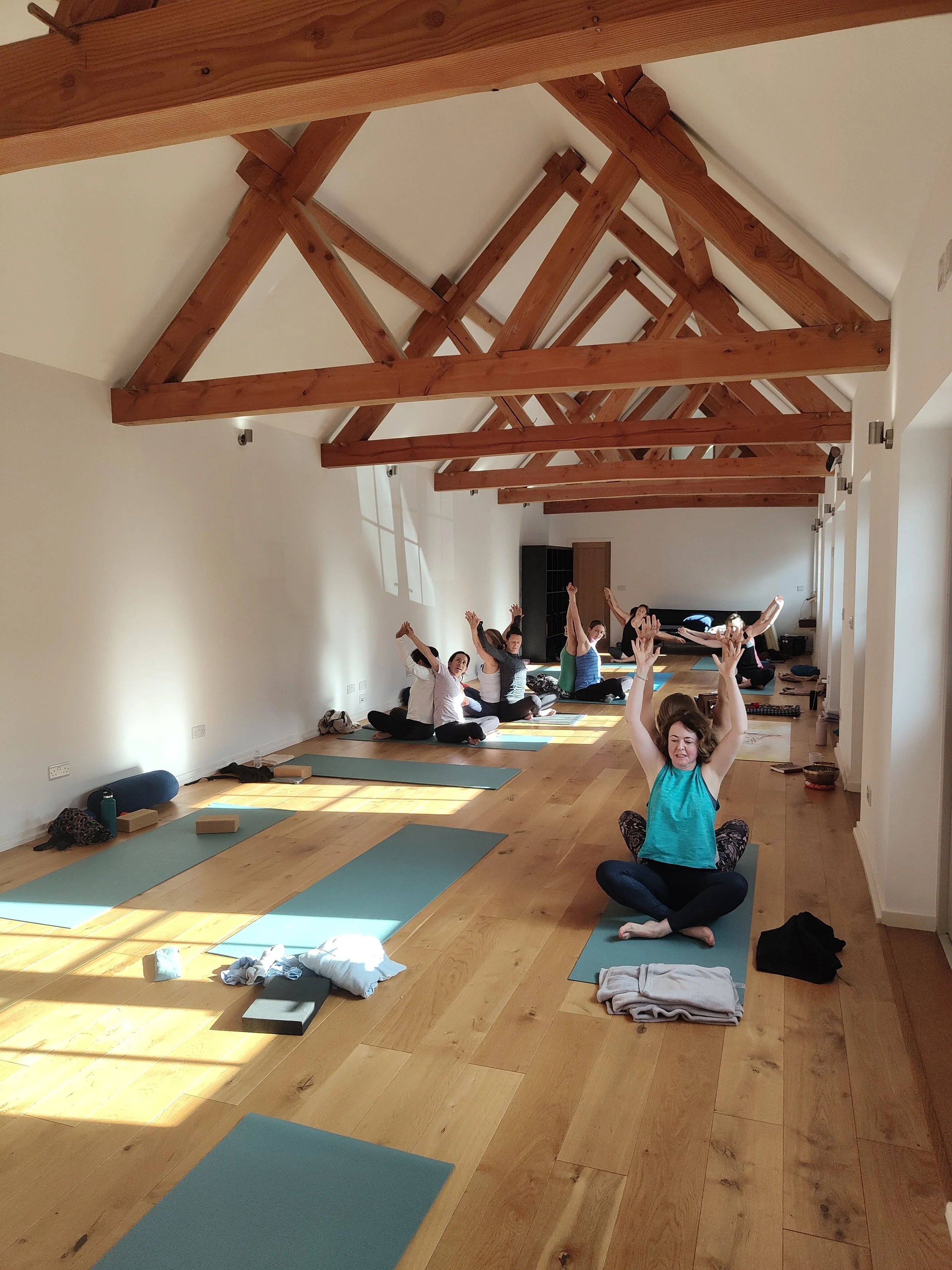 Group of people participating in a yoga or meditation class in a spacious studio with wooden beams and sunlight coming through windows.