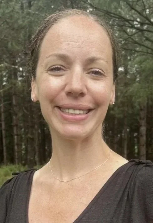 Close-up photo of a smiling woman with light skin and brown hair tied back, outdoors with trees in the background.