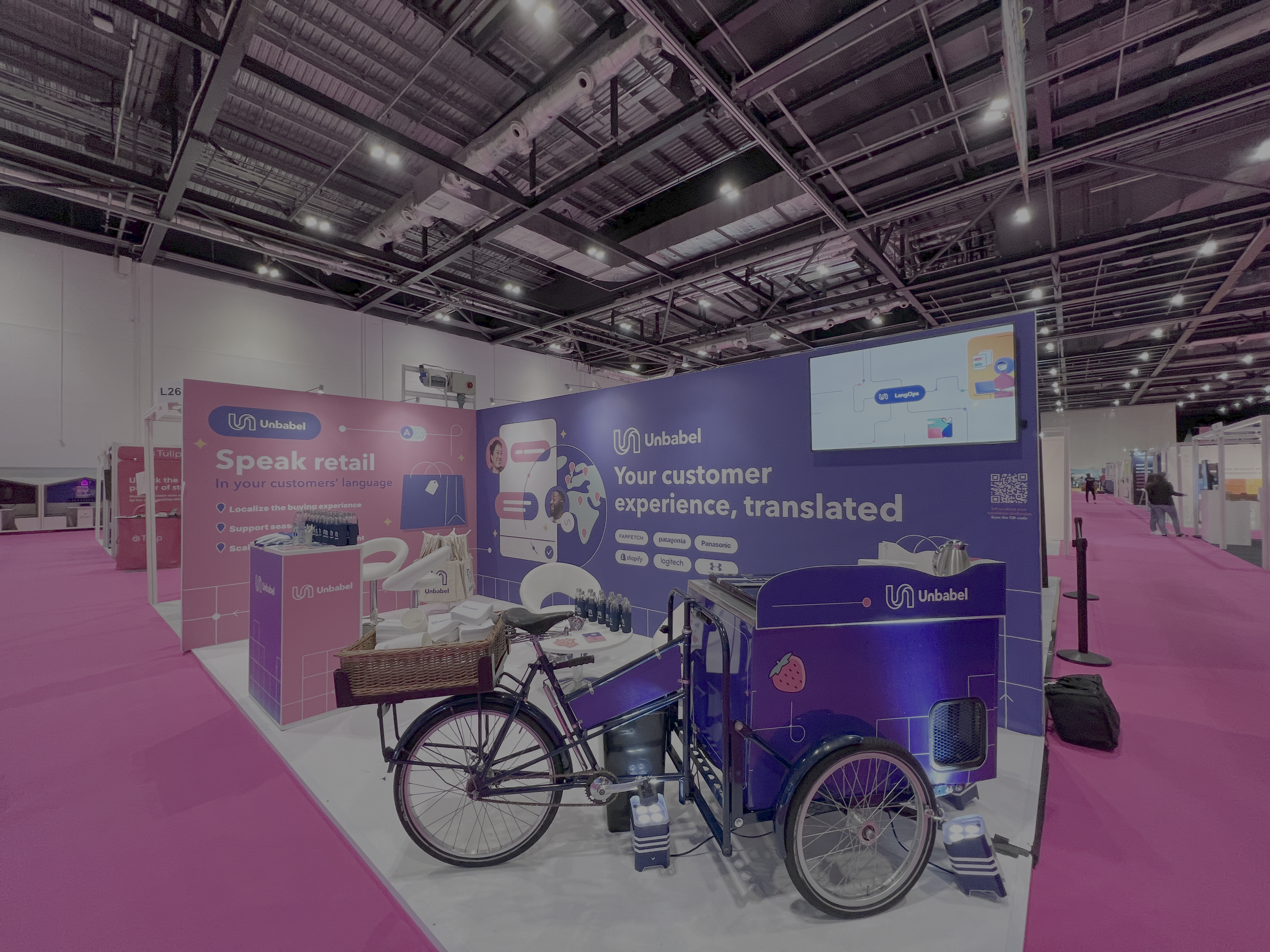 Trade show booth with purple and pink branding for Unbabel, featuring a bicycle with a cargo compartment, a monitor, and promotional materials.