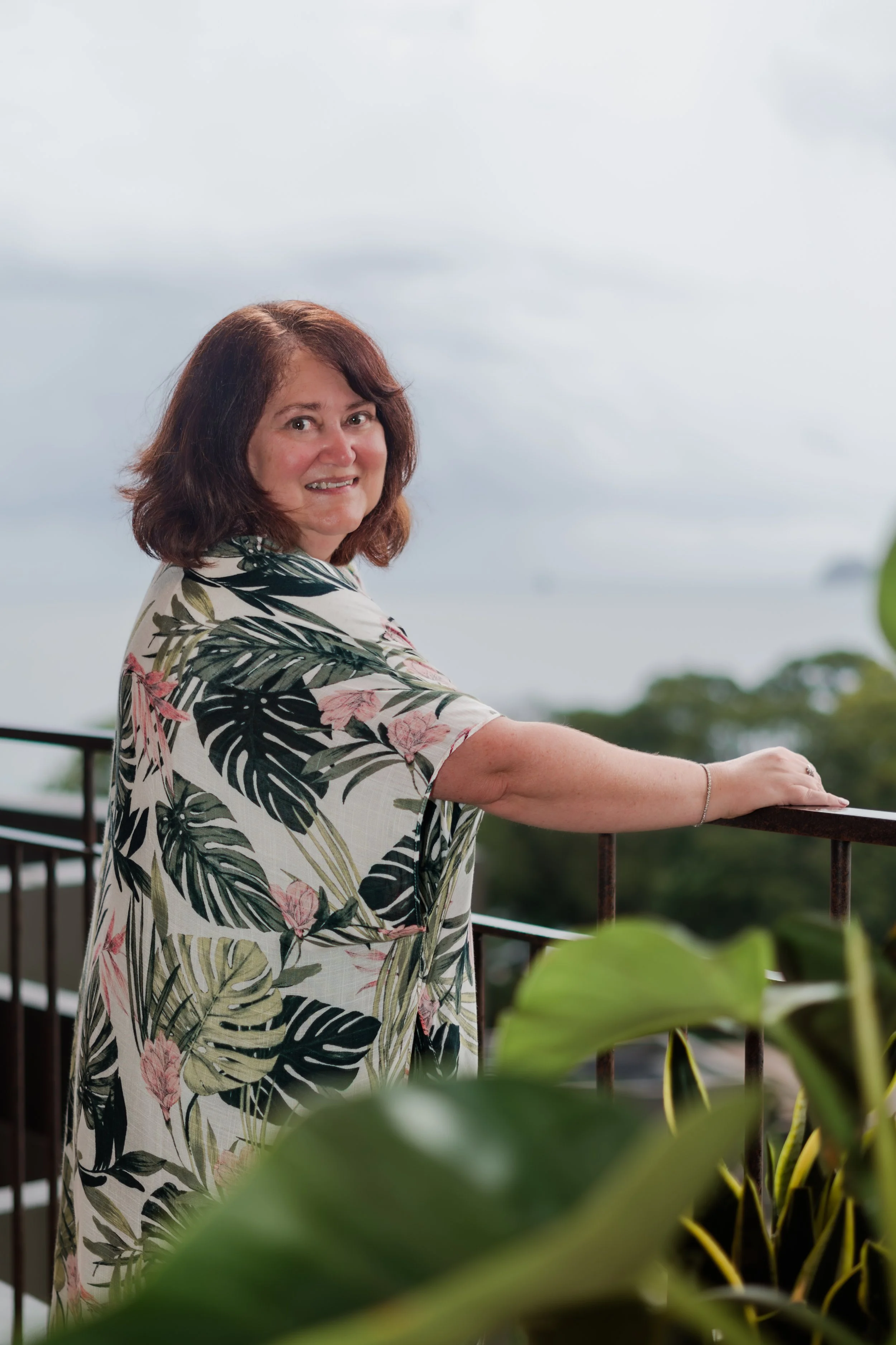 A woman with shoulder-length brown hair, smiling and leaning on a railing, wearing a tropical-patterned dress with green leaves and pink flowers, outdoors on a cloudy day with trees in the background.