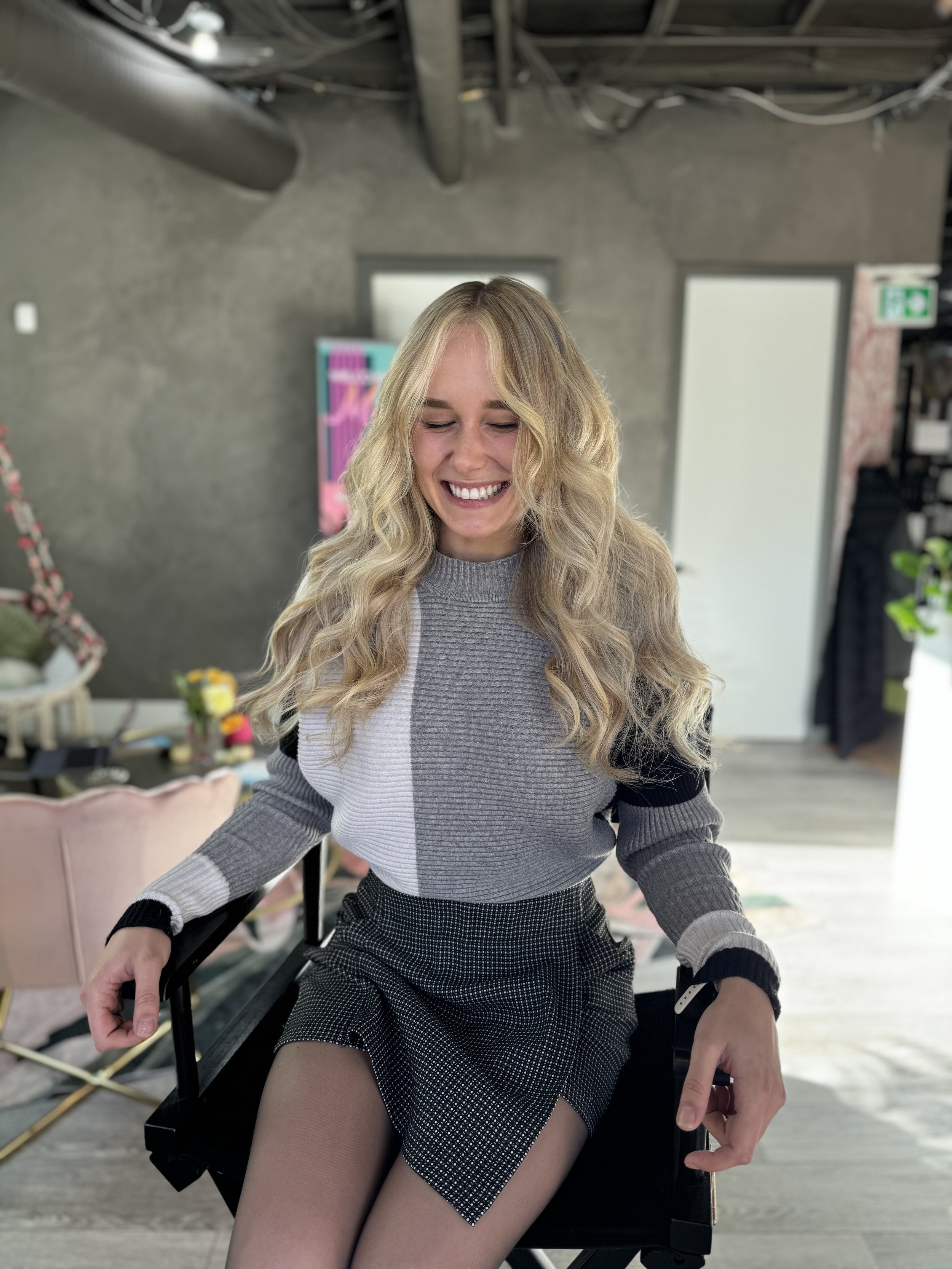 A young woman with long blonde curly hair sitting on a black chair, smiling with her eyes closed in a modern indoor space.
