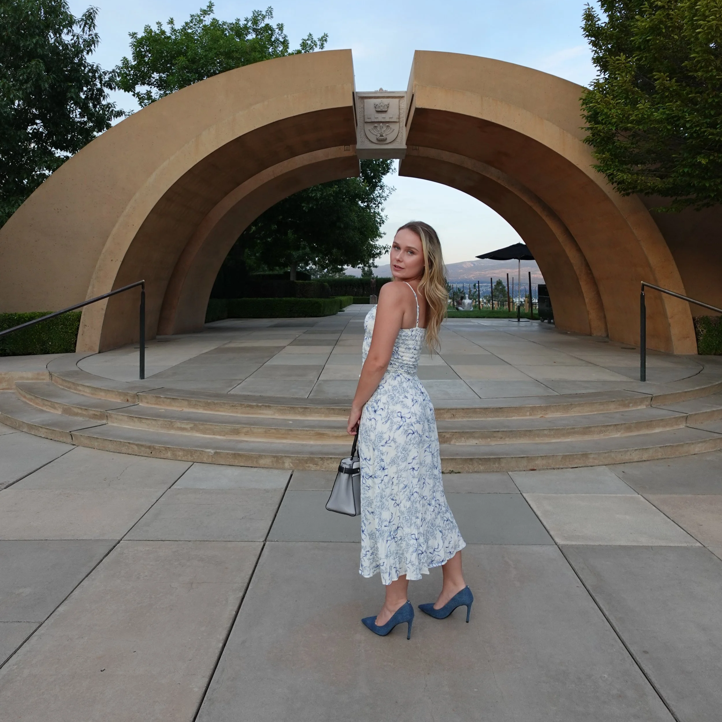 Woman in a white and blue floral dress, blue high heels, and carrying a small handbag, standing outdoors on a paved area in front of a large stone arch monument with steps.