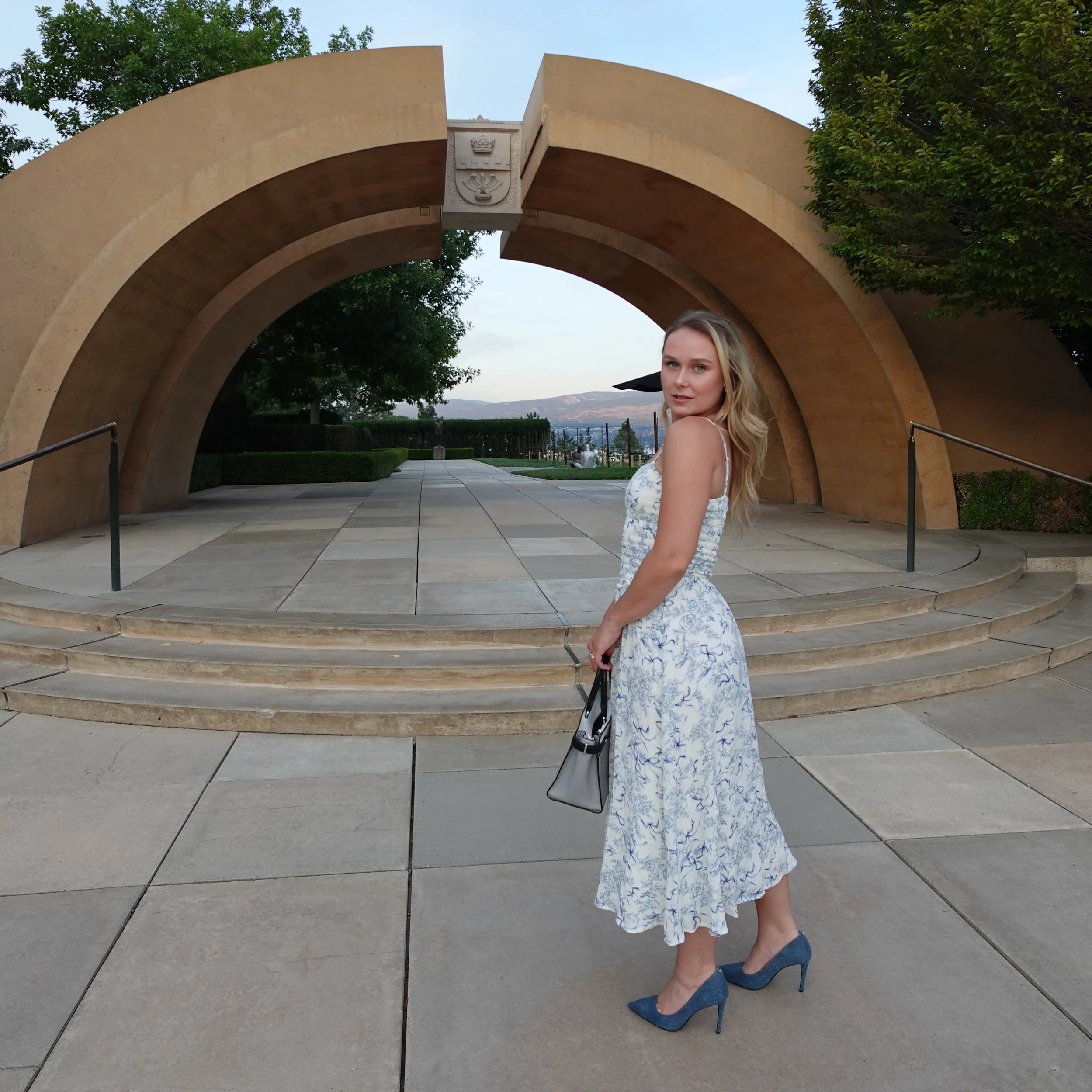 A woman in a white and blue floral dress with blue high heels holding a handbag, standing in front of a large stone arch structure during the daytime.