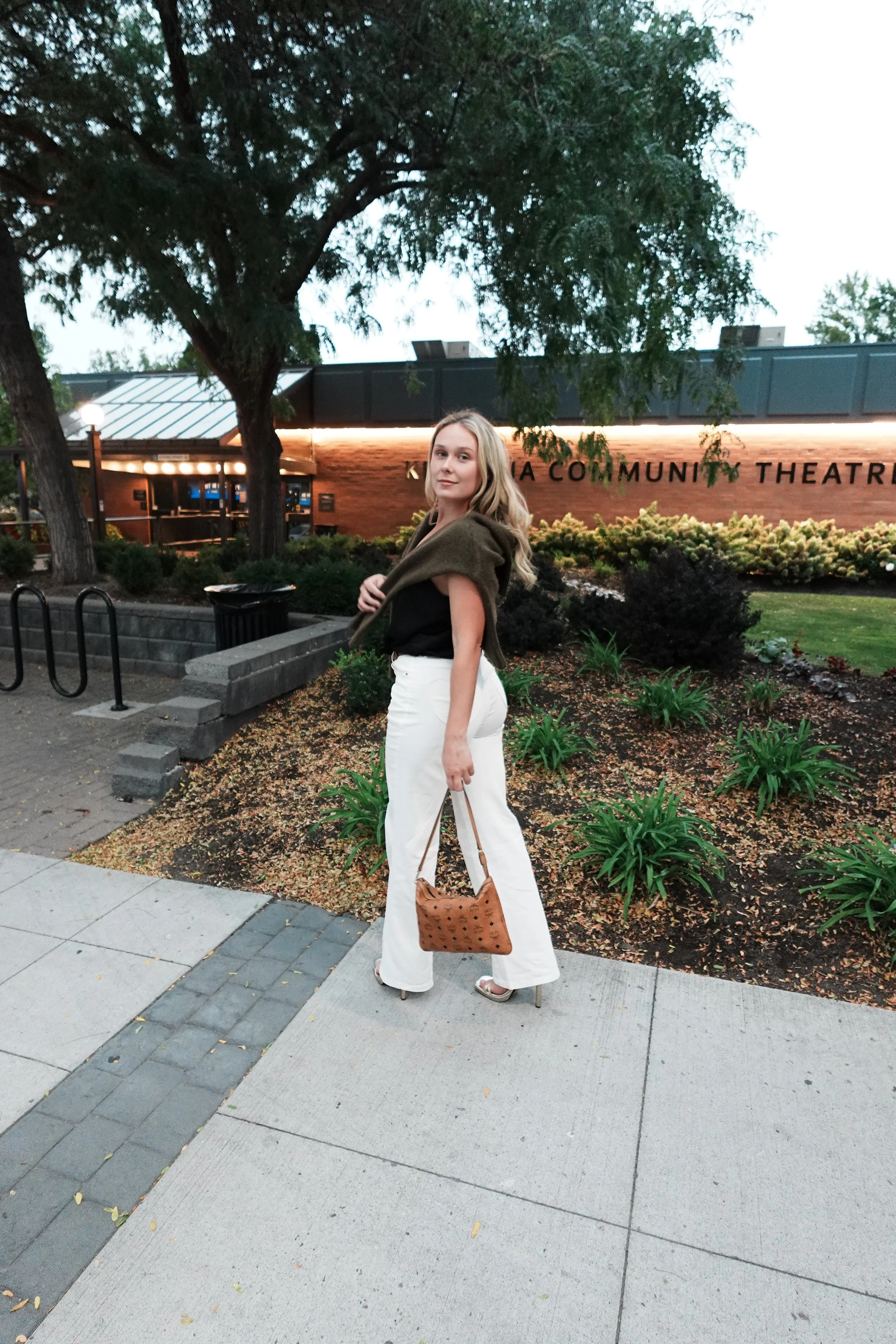 A woman stands on a sidewalk in front of a building with a sign that reads 'A COMMUNITY THEATRE'. She is wearing white pants, a black top, a brown jacket draped over her shoulders, metallic high heels, and is holding a brown purse. She has blonde hair and is posing with a slight smile.
