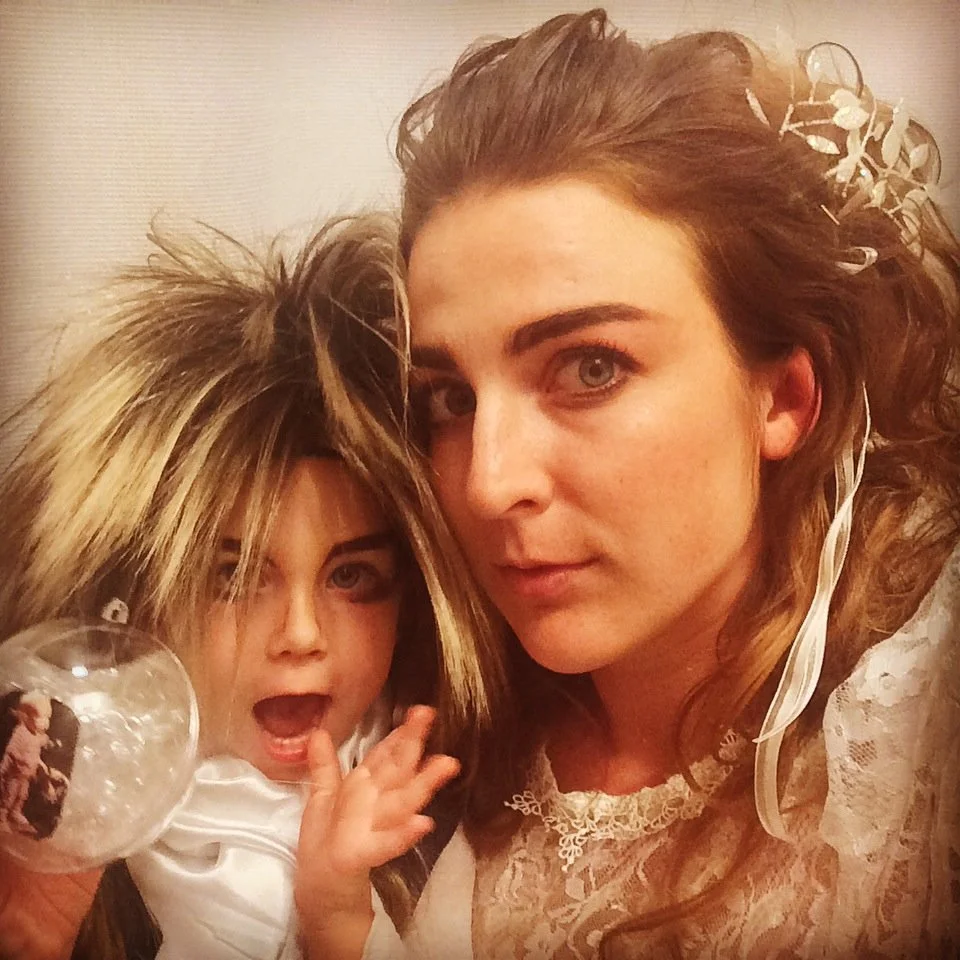 A woman and a young girl wearing long, messy blonde wigs, posing closely together. The woman has long, curly reddish-brown hair with a transparent floral hair accessory, and is wearing a lace top. The girl has her mouth open and is raising her hand, holding a glass with a photo on it, and appears to be surprised or excited.